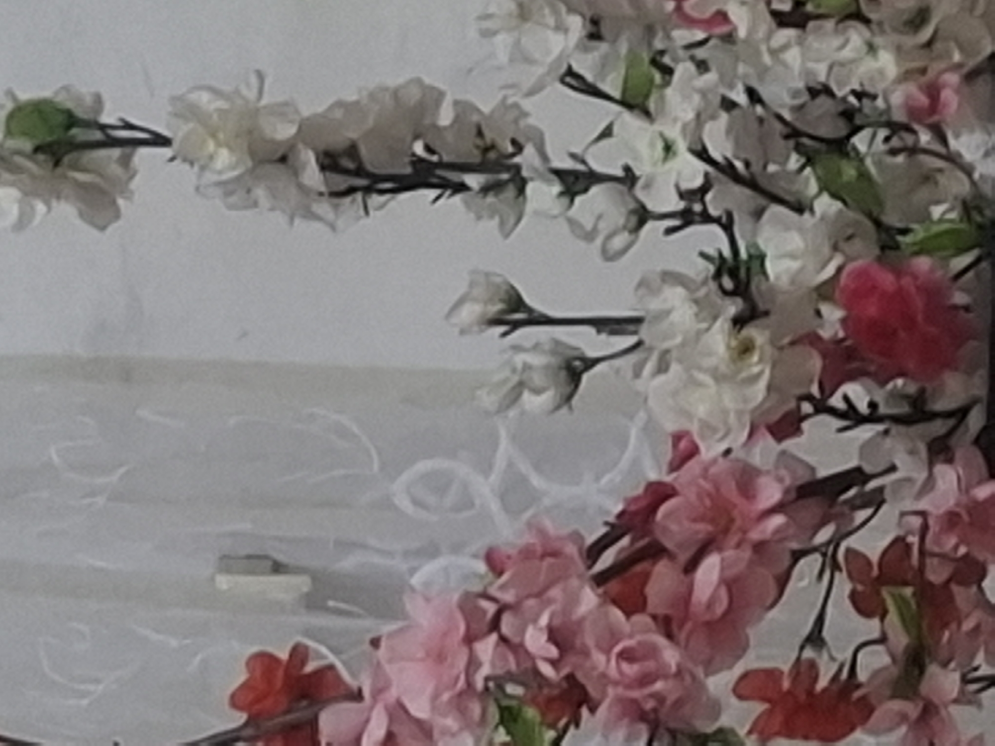 The image displays a close-up of artificial flowers, predominantly in shades of pink and white, arranged in a bouquet. The flowers are intricately detailed, with delicate petals and visible stems. In the background, a sheer white curtain with a subtle pattern is visible, suggesting an indoor setting. The lighting is soft, casting a gentle glow on the floral arrangement. The scene is serene and decorative, likely intended to enhance the ambiance of a room. The image does not contain any elements or contextual clues that would place it specifically in Almaty, Kazakhstan, or any other particular city.