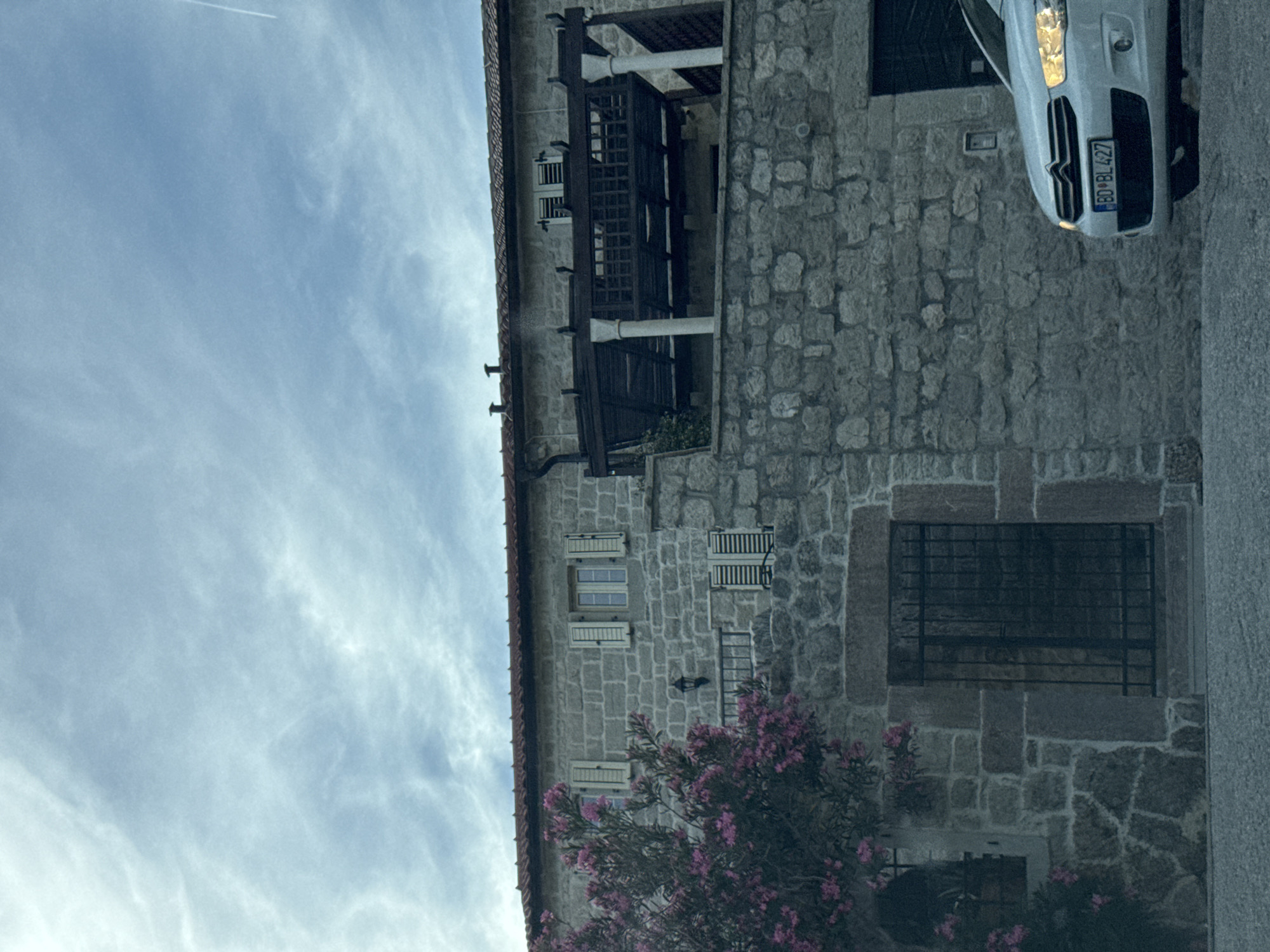 In the city of Rijeka Reževići, Montenegro, a white car with license plate BO BL 427 is parked in front of a rustic stone building. The car is facing towards the right of the frame, with its headlights on. The building features stone walls, wooden shutters on the windows, and a tiled roof. A flowering bush with pink blossoms is visible to the left of the building. The sky above is overcast with wispy clouds.