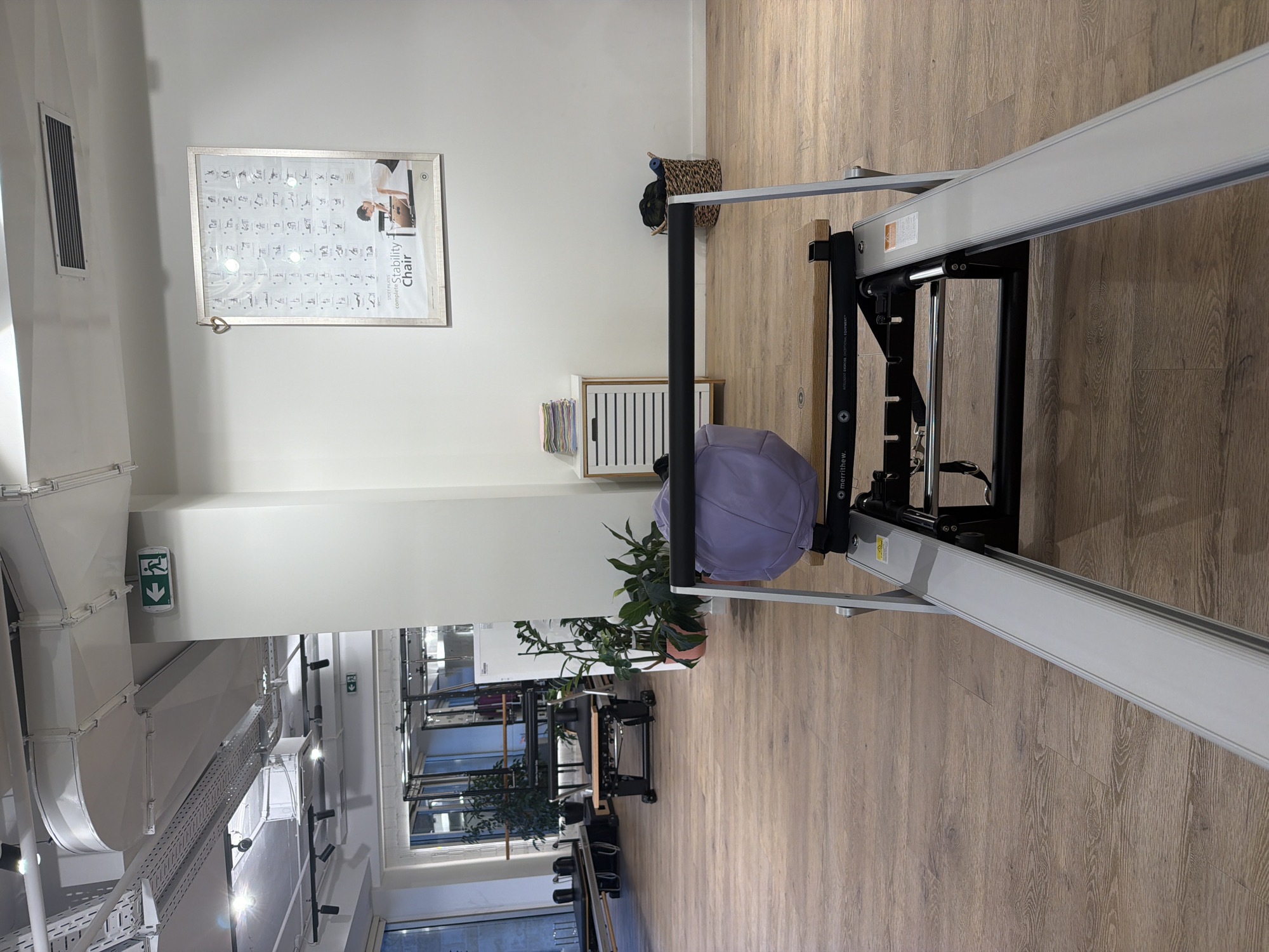 An indoor view of a Pilates studio, likely in Boreti, Montenegro, shows a Pilates reformer in the foreground. The reformer is metallic with a wooden seat and a light purple cushion. In the midground, a white cabinet with striped drawers holds folded colorful towels, and a wicker basket contains dark items. Beyond the cabinet, lush green plants in pots add a touch of nature. The studio floor is made of light-colored wood planks. In the background, other Pilates equipment and a large window are visible. The walls are painted white, and a framed poster displaying Pilates exercises hangs on the wall, featuring a diagram of the "STOTT PILATES complete Stability Chair." An illuminated green exit sign with a left-pointing arrow is mounted on the wall near the ceiling. The ceiling features exposed ductwork and lighting fixtures.