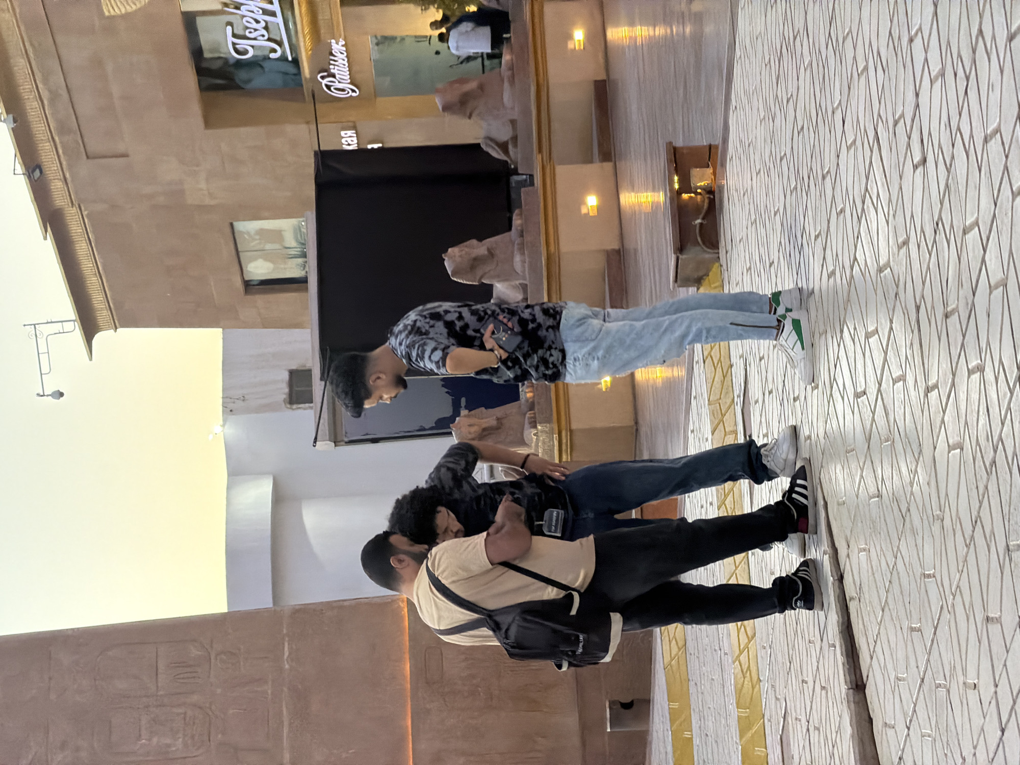 The image depicts a scene at the Sharm El Sheikh, Egypt, possibly within a themed shopping area. In the foreground, three young men are interacting. Two men on the left are looking at a phone, with one wearing a beige t-shirt and black pants, carrying a black backpack. The other man, in the center, has curly dark hair and is wearing a dark, patterned t-shirt and blue jeans. To the right, a third man, wearing a similar dark, patterned t-shirt and light blue jeans, stands facing left with his hands on his hips.
The environment features a paved courtyard with a geometric tile pattern. In the background, there are Egyptian-themed architectural elements, including a large obelisk with hieroglyphs on the left and sphinx statues in front of a building on the right. The building has a tan facade with windows and signage, one of which reads "Patisser" and another appears to say "Tseph". The sky is a pale yellow, suggesting either sunrise or sunset. Lighting fixtures are visible on the building and on poles, and illuminated square elements are embedded in a low wall. A security camera is mounted on a pole in the upper right portion of the image.
