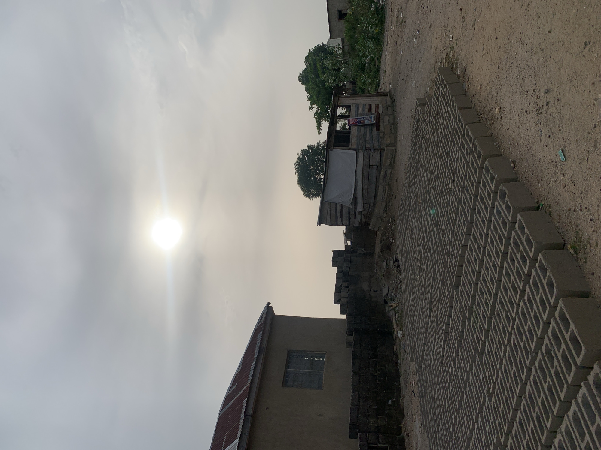 The scene depicts a building site on the outskirts of Adankolo, Nigeria, in the early morning or late afternoon. The sky is overcast, with the sun obscured by clouds. In the foreground, stacks of grey concrete blocks are neatly arranged. A small, makeshift wooden structure stands nearby, possibly a storage shed or a vendor's stall. A single-story building with a corrugated metal roof is also visible. The ground is dusty and barren. No people are visible in the frame.
