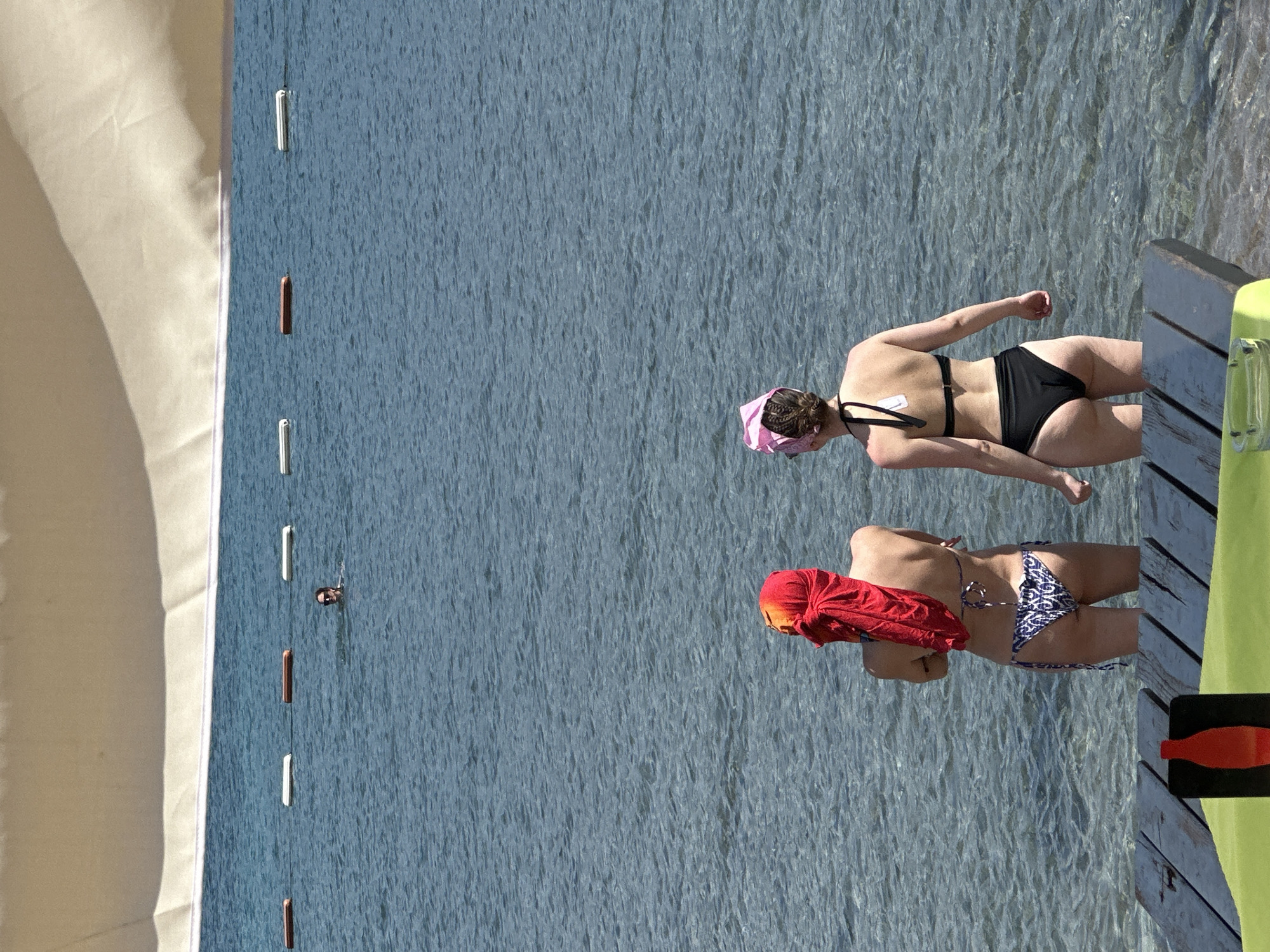 The image shows a beach scene in Budva, Montenegro. Two women in bikinis are standing on a wooden pier extending into the water. One woman is wearing a red headscarf and a patterned bikini, while the other is wearing a pink headscarf and a black bikini. In the background, there's a white sunshade and buoys marking a swimming area in the sea. A swimmer is also visible in the water. Part of a green paddleboard or similar watercraft is visible on the pier.
