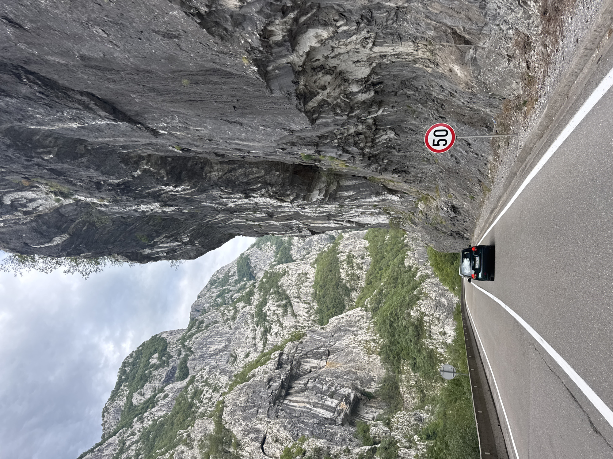 The image shows a road passing through a narrow mountain gorge. A dark-colored car is driving on the road, heading away from the camera. To the right of the road, there is a speed limit sign indicating 50. The surrounding environment is dominated by steep, rocky mountain slopes covered with vegetation. The sky is overcast. The image is taken from a moving vehicle. This scene is located on the road to the Skadar Lake National Park, near Vlahovići, Montenegro.
