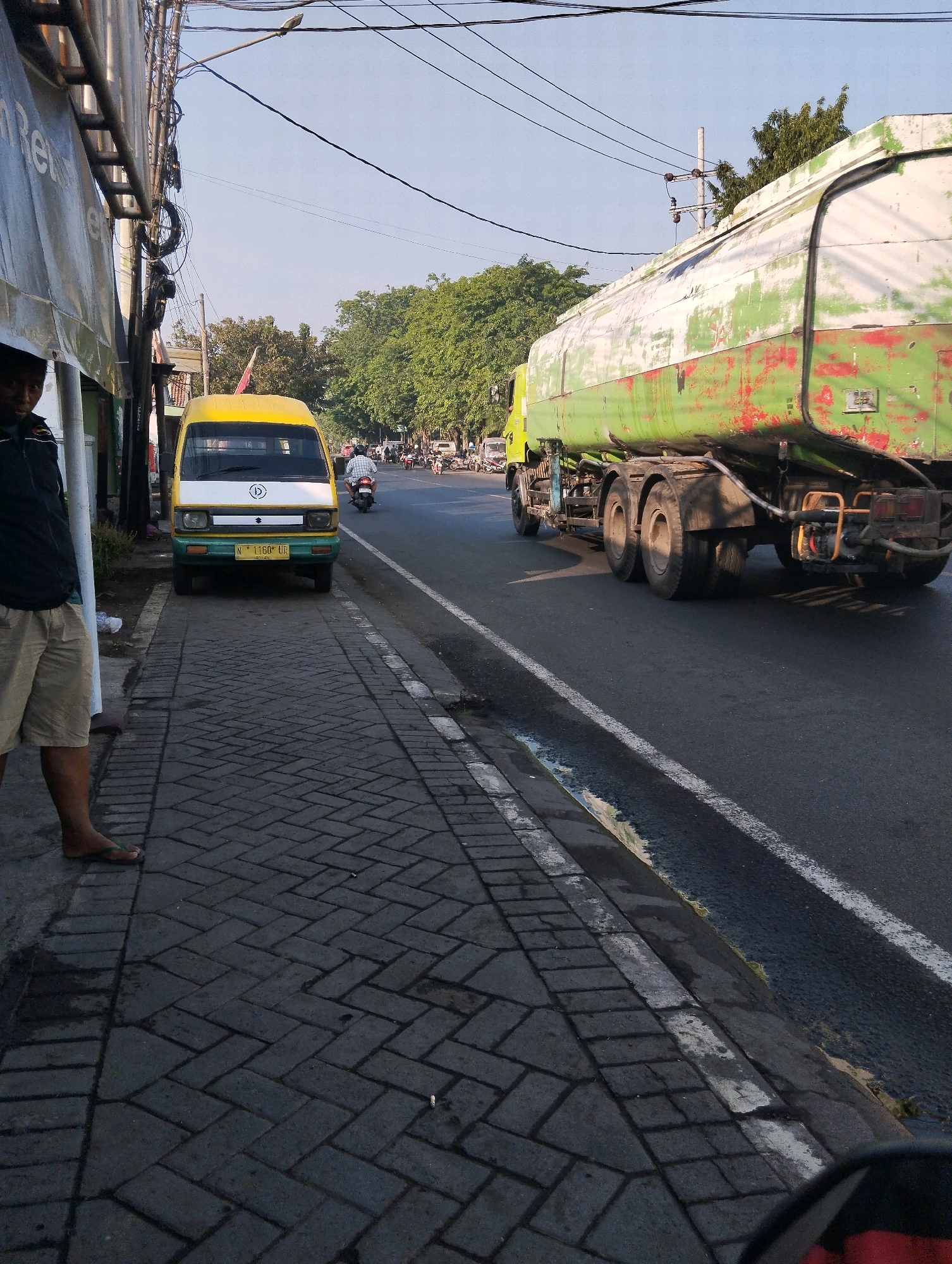 The image depicts a street scene in Probolinggo, Indonesia. On the left, a person stands on a paved sidewalk next to a parked yellow and white van. The van has a license plate that reads "N 1160 UR". Further down the street, a large green and white tanker truck is in motion on the asphalt road. The tanker truck appears weathered, with patches of red visible beneath the green paint. The street is lined with trees on the right side. The sky is visible in the background, with overhead power lines crisscrossing the frame. The overall impression is a typical urban street with traffic and pedestrians.