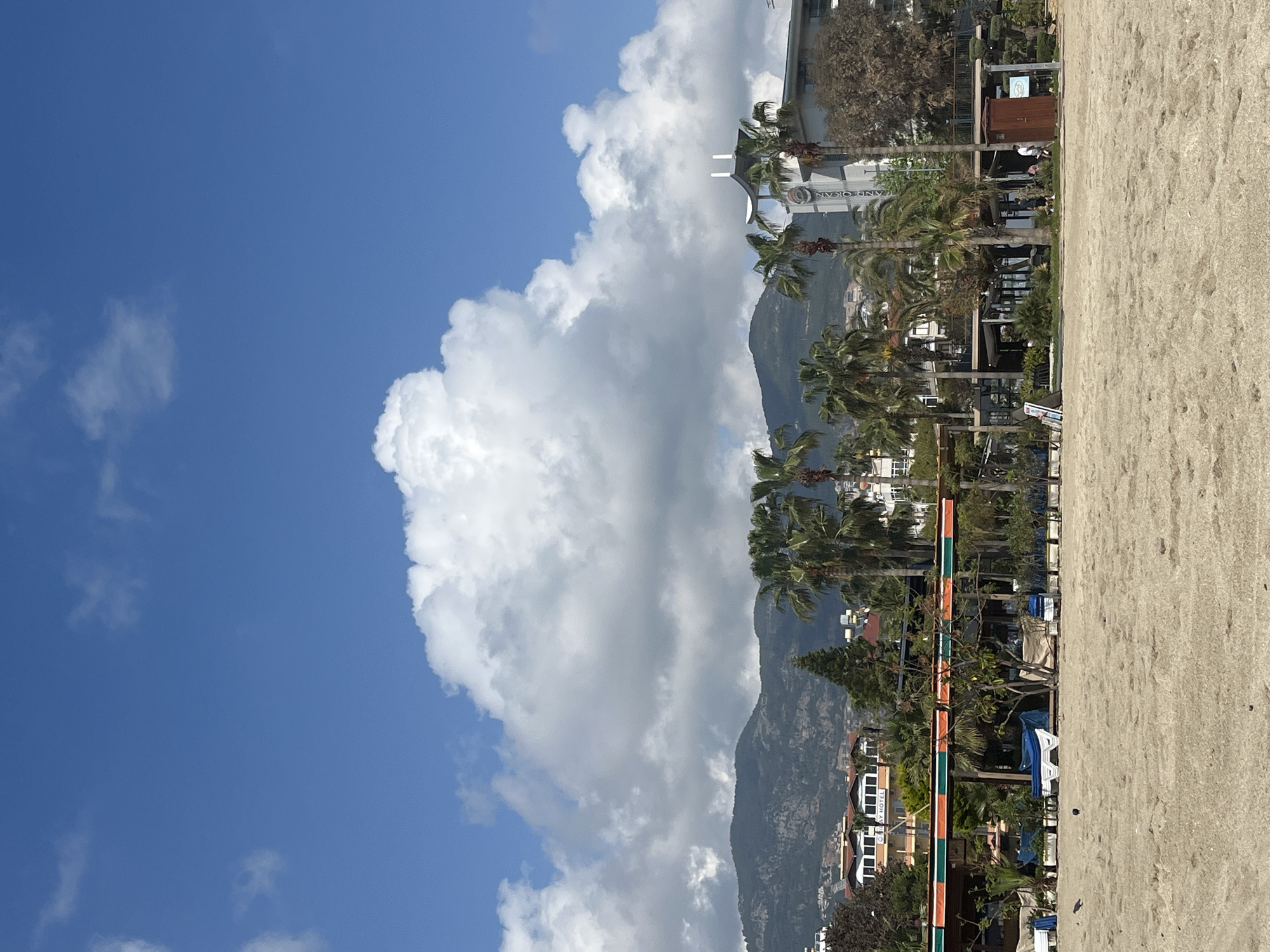 The scene is set on a beach in Alanya, Turkey, with a view of the Taurus Mountains in the background. The sky is partially covered with large cumulus clouds. In the midground are palm trees and various buildings. Beach sand occupies the right side of the frame. There are very distant people, but they are difficult to distinguish.