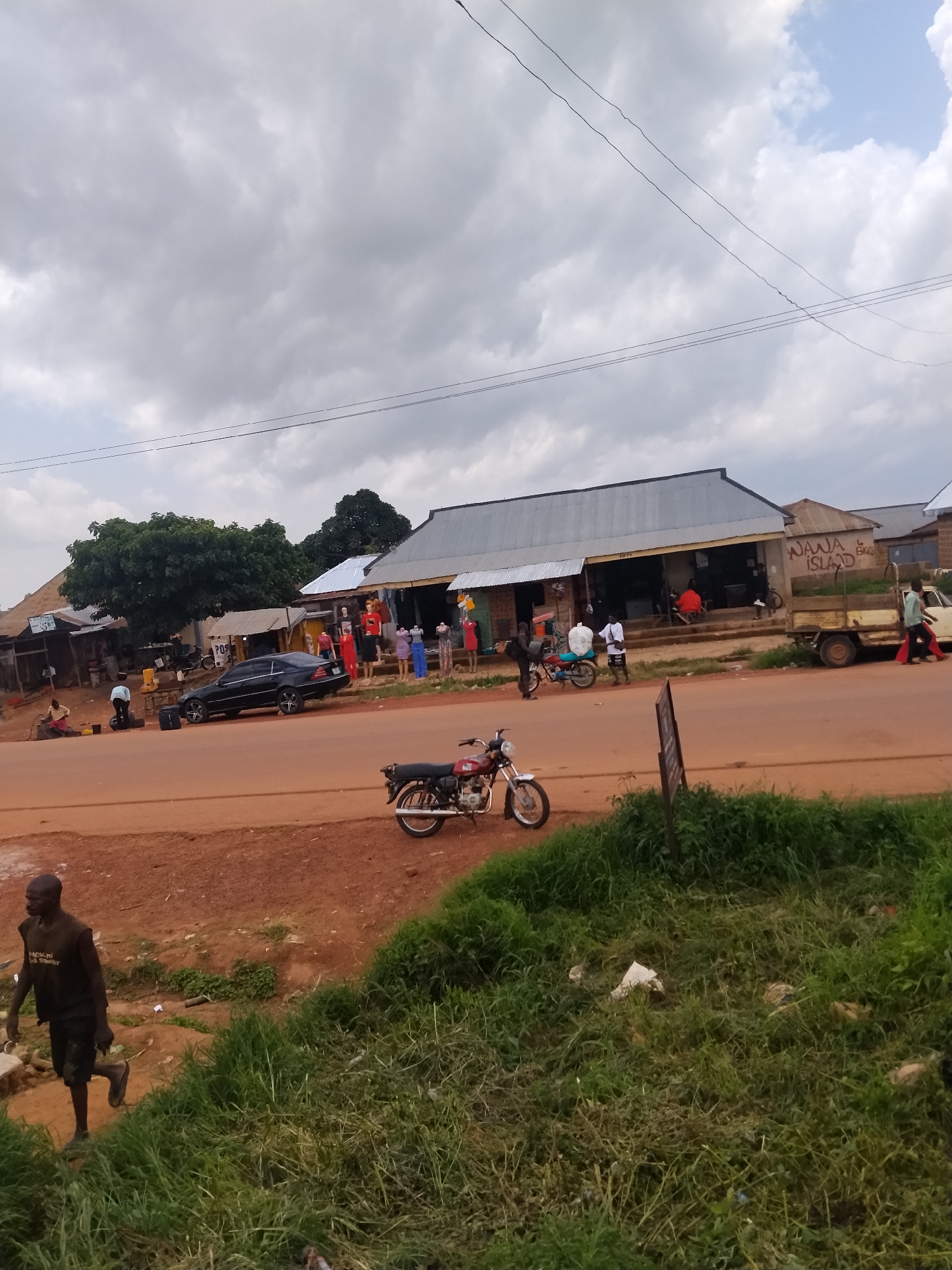 The image depicts a street scene in Durumi, Nigeria. A dirt road runs horizontally across the foreground, bordered by patchy green grass and some debris on the right. A man in a black t-shirt and shorts walks away from the camera on the left side of the road.
Across the road, a row of single-story buildings with corrugated metal roofs lines the street. One building has the words "NAWA ISLAND" painted on its facade. Several mannequins dressed in colorful clothing are displayed outside one of the shops. A black sedan is parked in front of these buildings. A few people are visible in the vicinity of the shops, some interacting near motorcycles and a flatbed truck. The sky is overcast with scattered clouds. The overall impression is a bustling, informal commercial area.