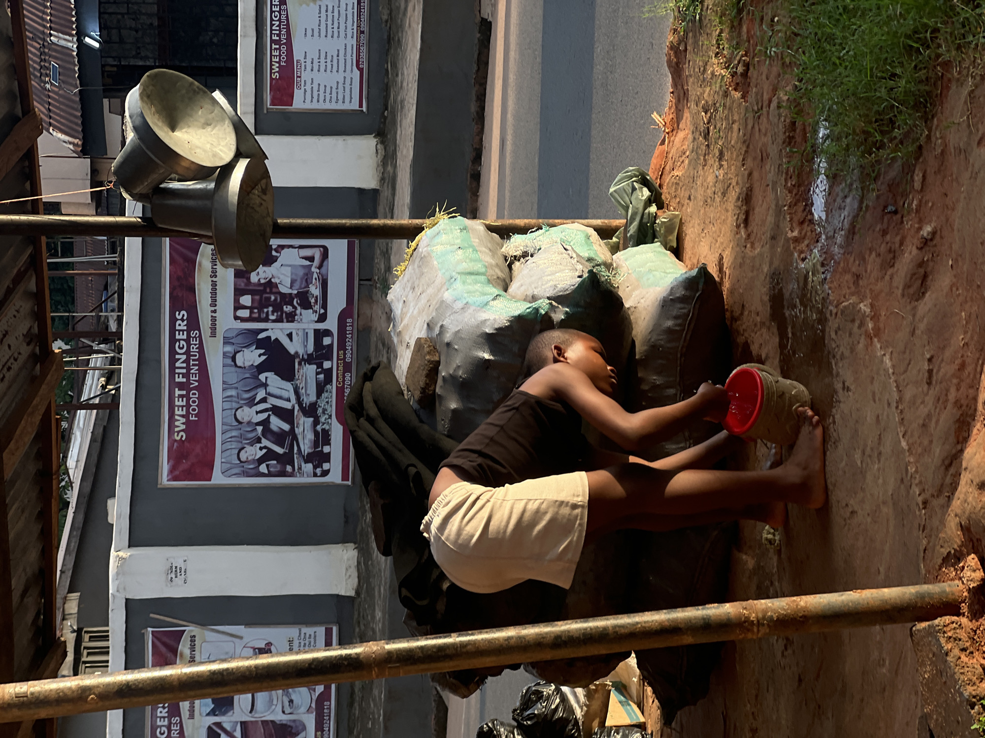 A young child, likely a boy, is shown in a full-body shot, lying on his stomach with his legs outstretched. He is wearing a dark tank top and light-colored shorts. The child is holding a red and white plastic container, from which he is pouring water onto a pile of sacks. The sacks appear to be made of rough material, some light green and others dark, and are stacked on the ground. To the left of the child, there are some metal pots hanging.
The scene is outdoors, with the ground covered in reddish-brown dirt and patches of green grass visible further up. A paved road is visible in the background, suggesting an urban or semi-urban setting. There are also posters on a building behind the sacks. One poster reads "SWEET FINGERS FOOD VENTURES" and lists menu items. Another poster, partially visible, also mentions "SWEET FINGERS". The lighting suggests it is daytime, possibly late afternoon, given the shadows. The child's posture and the activity of pouring water imply he might be tending to something or engaged in some form of play or chore. The overall impression is of a candid, everyday moment captured in a humble setting. The text on the posters indicates a business named "Sweet Fingers Food Ventures" in Nkpor, Nigeria, specializing in food services and possibly indoor/outdoor services.