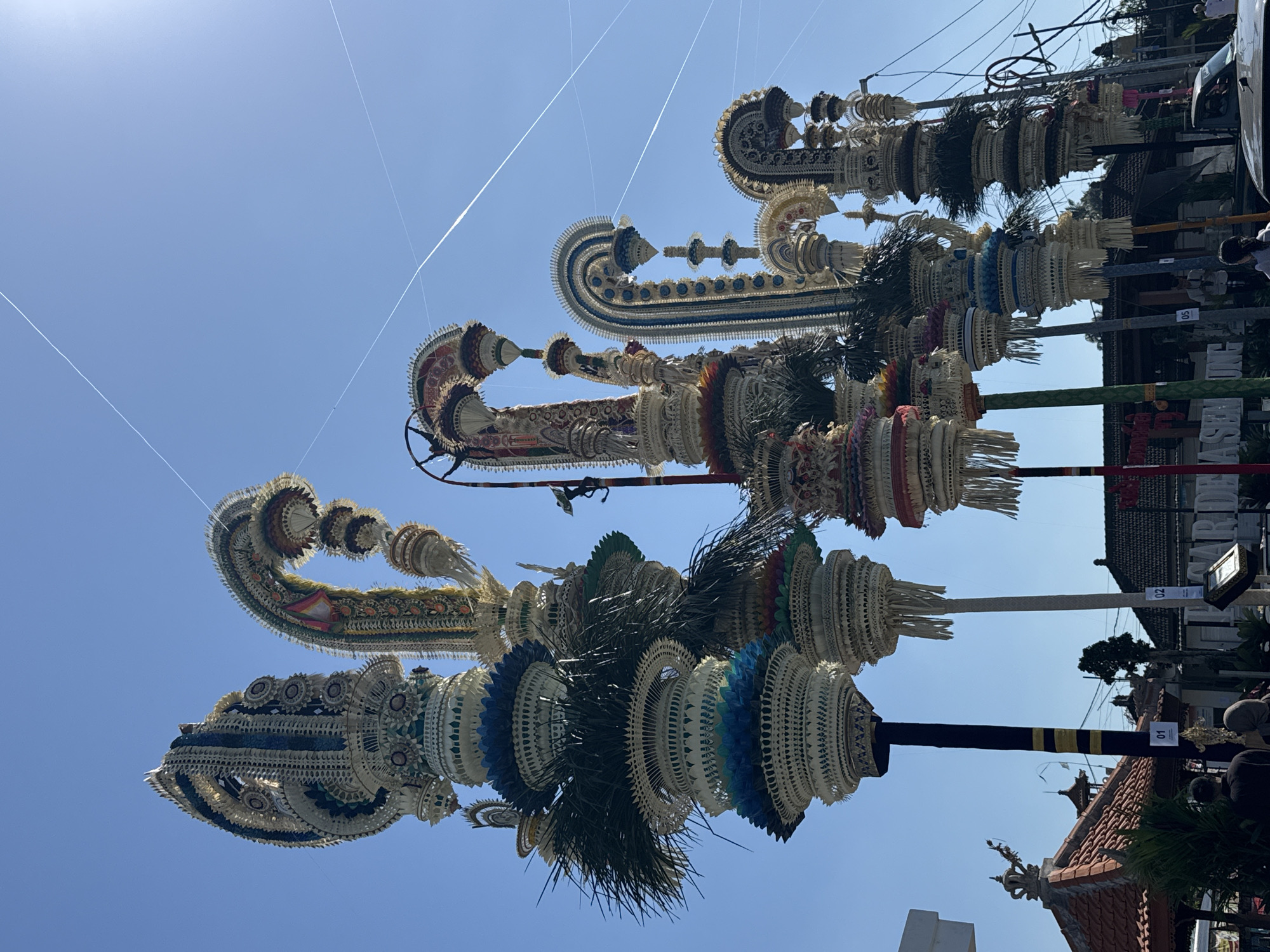 The image depicts a bright, sunny day in Mengwi, Indonesia, characterized by a clear blue sky with thin white contrails from aircraft. The foreground is dominated by a line of tall, elaborately decorated ceremonial structures, likely for a festival or religious event. These structures are made of woven natural materials, primarily palm leaves, and are adorned with intricate patterns, colorful beads, and dangling fringes. They vary in height and design, with some featuring curved, arching tops and others standing more upright. Several of these structures have small numbered signs at their bases, labeled "01," "02," and "05."
In the background, traditional Balinese architecture is visible, including buildings with red-tiled roofs and ornate carvings. A few people are present, appearing as silhouettes or blurred figures in the lower parts of the image, suggesting they are spectators or participants in the event. One person is seen from behind wearing a headscarf, and another is standing near a sign. A white vehicle is also partially visible on the right side of the frame. The overall impression is one of cultural festivity and community gathering under a clear sky.