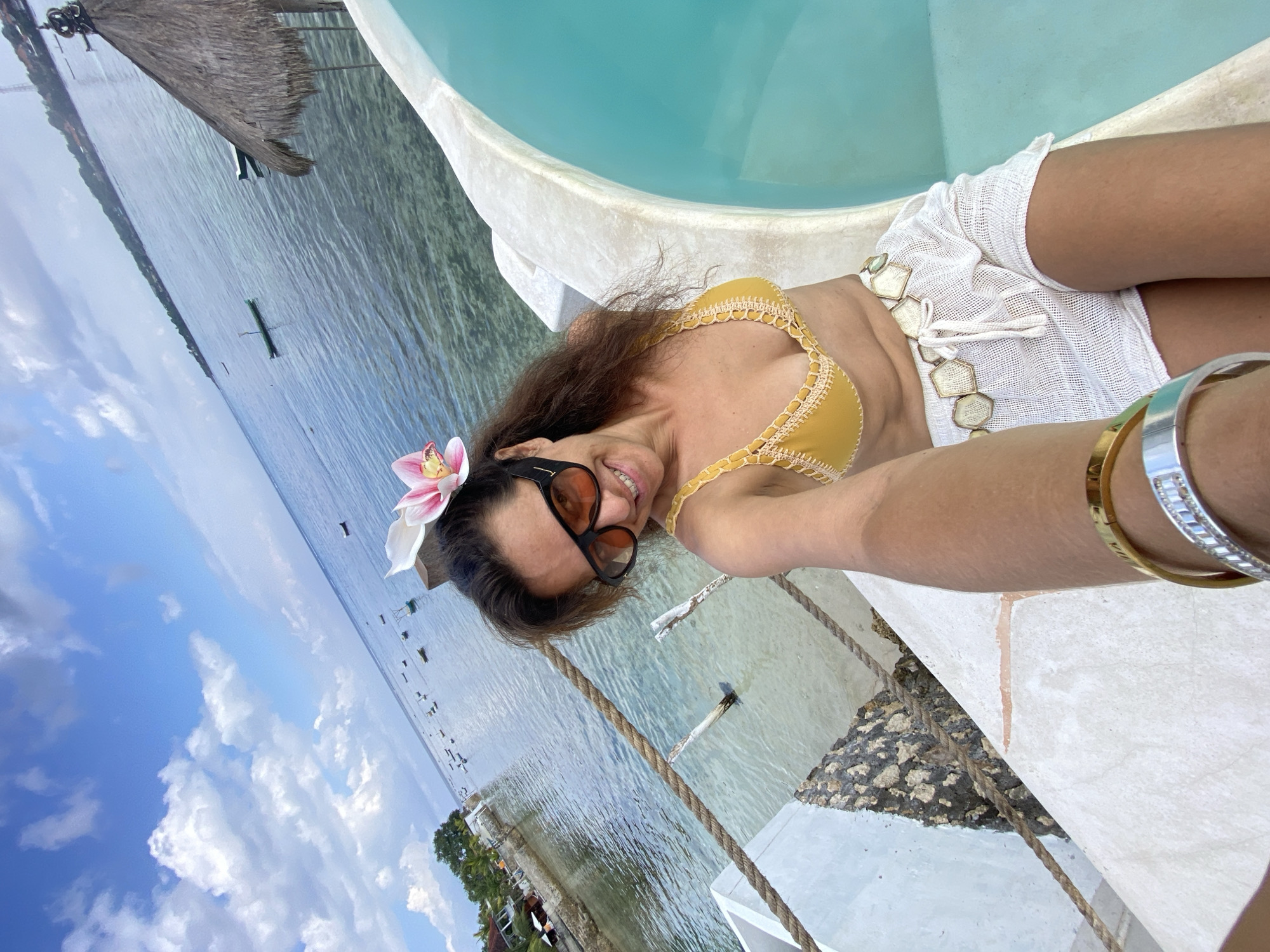 A woman is captured in a selfie by a pool overlooking the ocean in Nusa Penida, Indonesia. She is wearing a yellow bikini top, white shorts, sunglasses, and a pink orchid in her hair. The pool's turquoise water contrasts with the clear blue sky dotted with white clouds. In the background, numerous small boats are scattered across the calm sea, and the coastline of Nusa Penida, with its lush greenery and buildings, is visible. The scene evokes a relaxed, tropical vacation atmosphere.