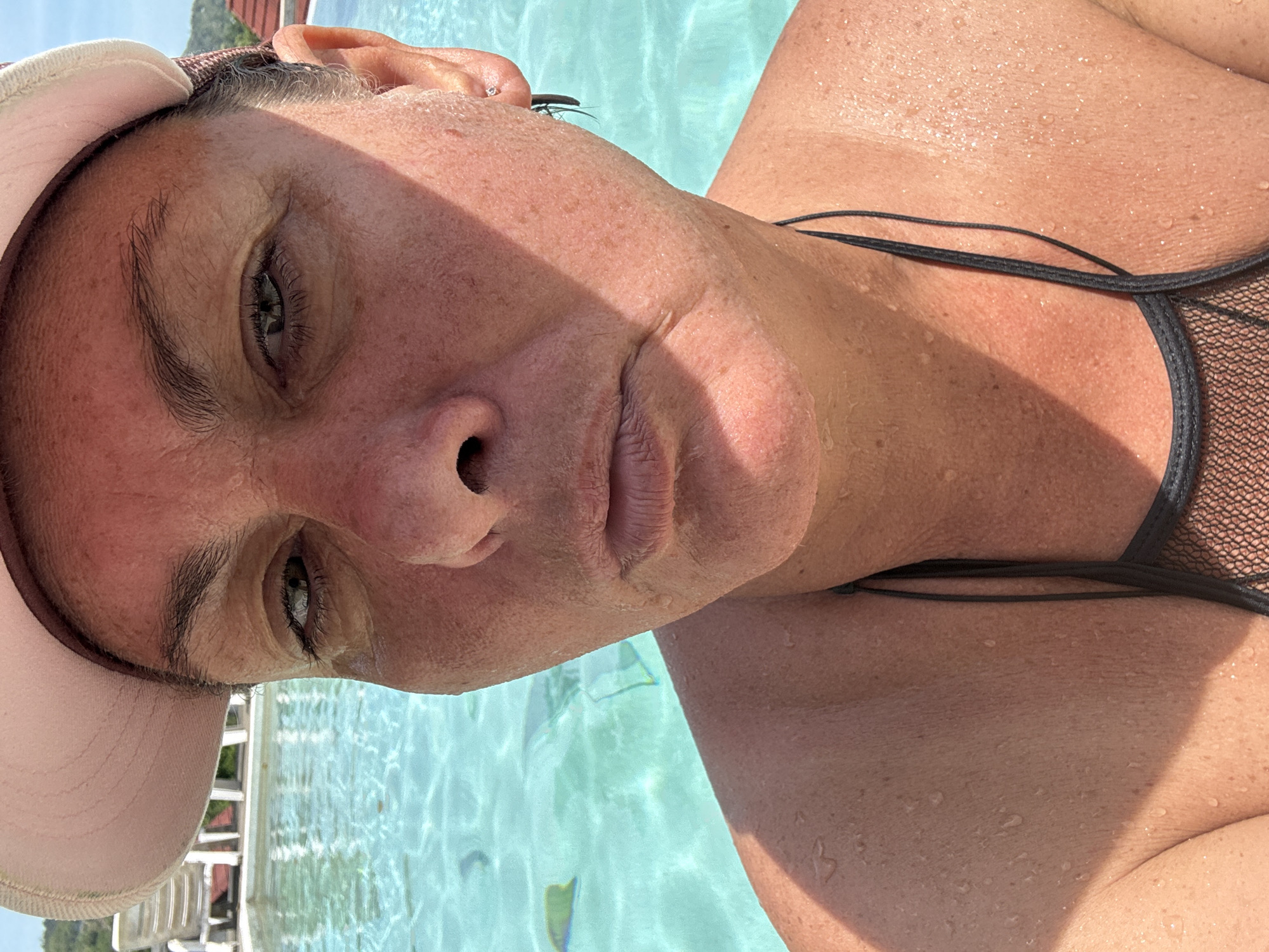 A close-up, slightly rotated image shows a fair-skinned person in a body of water, presumably swimming. The individual is wearing a pale pink baseball cap. A black bikini top is visible. Water droplets are scattered across the person's face and upper chest. The background shows turquoise water and the side of a structure. It is not possible to verify the specific location of บ้านบางรักษ์, Thailand without additional contextual information.
