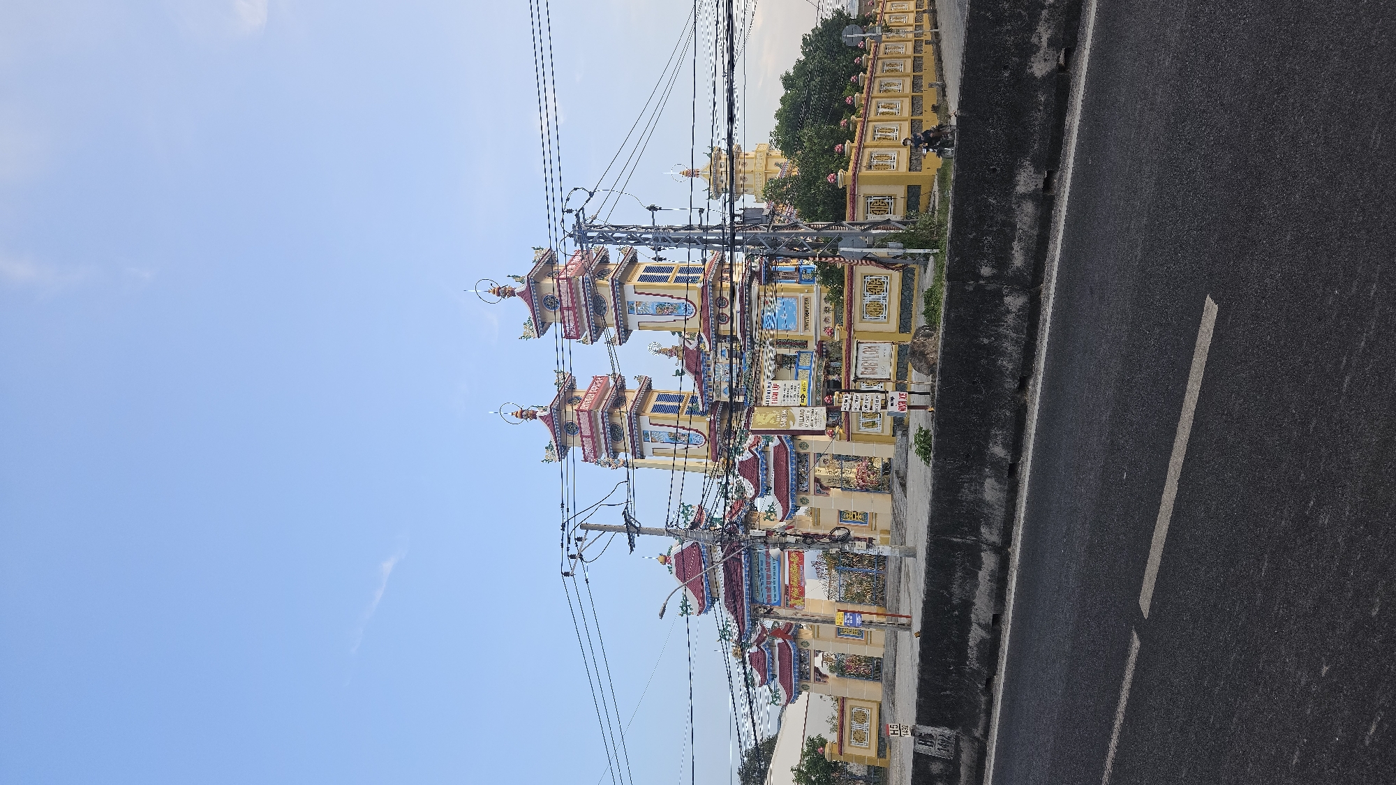 The image depicts a view alongside a roadway in Cam Đức, Vietnam. A series of brightly colored, tiered structures akin to pagodas, possibly funerary monuments, are situated beside the road. The structures feature layered roofs, ornate decorations, and a yellowish base color, mixed with red and blue accents. Overhead, a web of electrical wires is strung along utility poles. Further in the background, additional similar-style buildings and trees can be seen. The road, marked with a white line, is asphalt. The sky is clear blue.