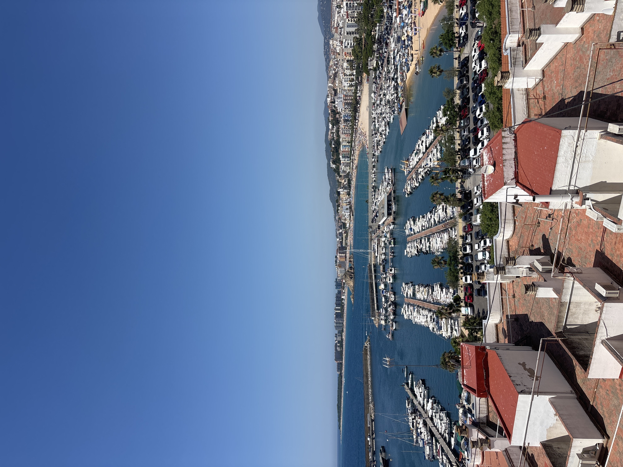 This is an aerial, eye-level, wide shot of a marina and coastline in Blanes, Spain. The foreground shows the rooftops of several white buildings with red tile roofs, some with chimneys and air conditioning units. Beyond the rooftops is a large marina filled with many docked white boats of various sizes. A road with parked cars separates the marina from a sandy beach with many people sunbathing and swimming in the blue ocean. Beyond the beach, a city skyline with numerous buildings, some taller than others, stretches along the coastline, with hills in the background. The sky is clear and blue.
