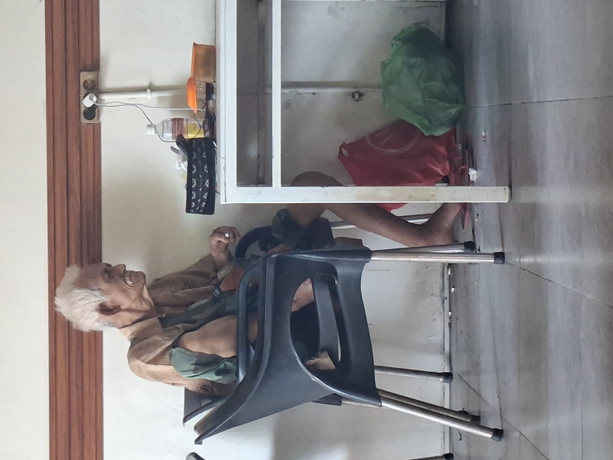 An elderly man sits on a black plastic chair, his legs extended under a white metal shelf. He is fair-skinned with short, white hair, and wears a tan or light brown shirt with a patterned green covering part of his chest and shoulder. He is turned toward the left side of the image, looking upwards with his mouth open. Under the shelf are several items, including a red bag and a green plastic bag. Above the shelf are various items, including bottles and containers. The scene is indoors, with tiled flooring and a wall that includes a wooden doorframe on the left side of the image. An electrical outlet and several connected wires are visible on the wall near the doorframe. There are no indications in the image to verify it being in Lhokseumawe, Indonesia.