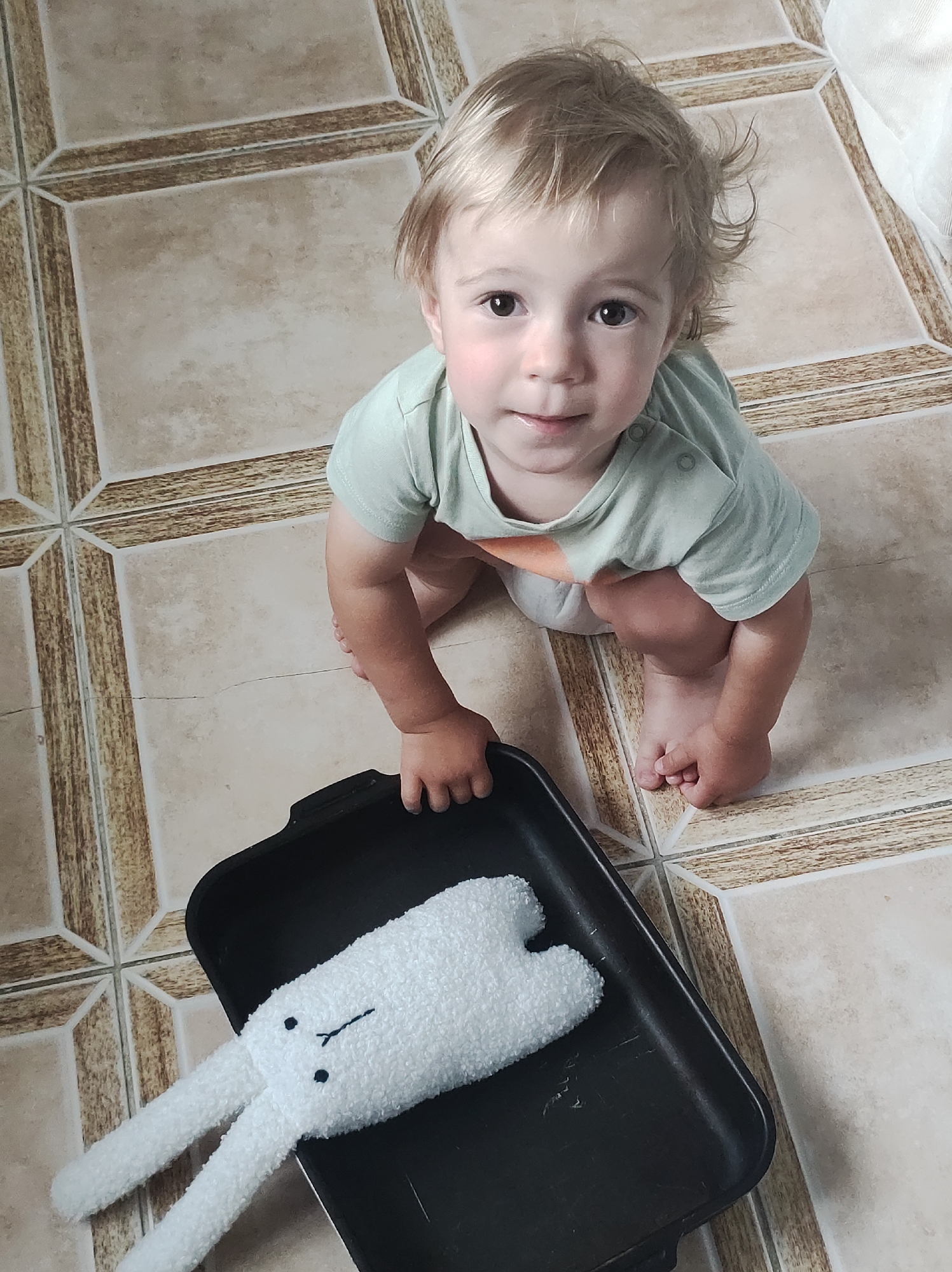 A young child with blonde hair is squatting on a beige tiled floor. The child is wearing a light green t-shirt and white diaper. In front of the child is a black tray containing a white stuffed rabbit toy. The child's left hand is resting on the edge of the tray. The background shows a continuation of the beige tiled floor, with darker grout lines forming a cross pattern. The image appears to be taken indoors. The image is not suitable for providing location information within the city of Chaikovsky, Russia.