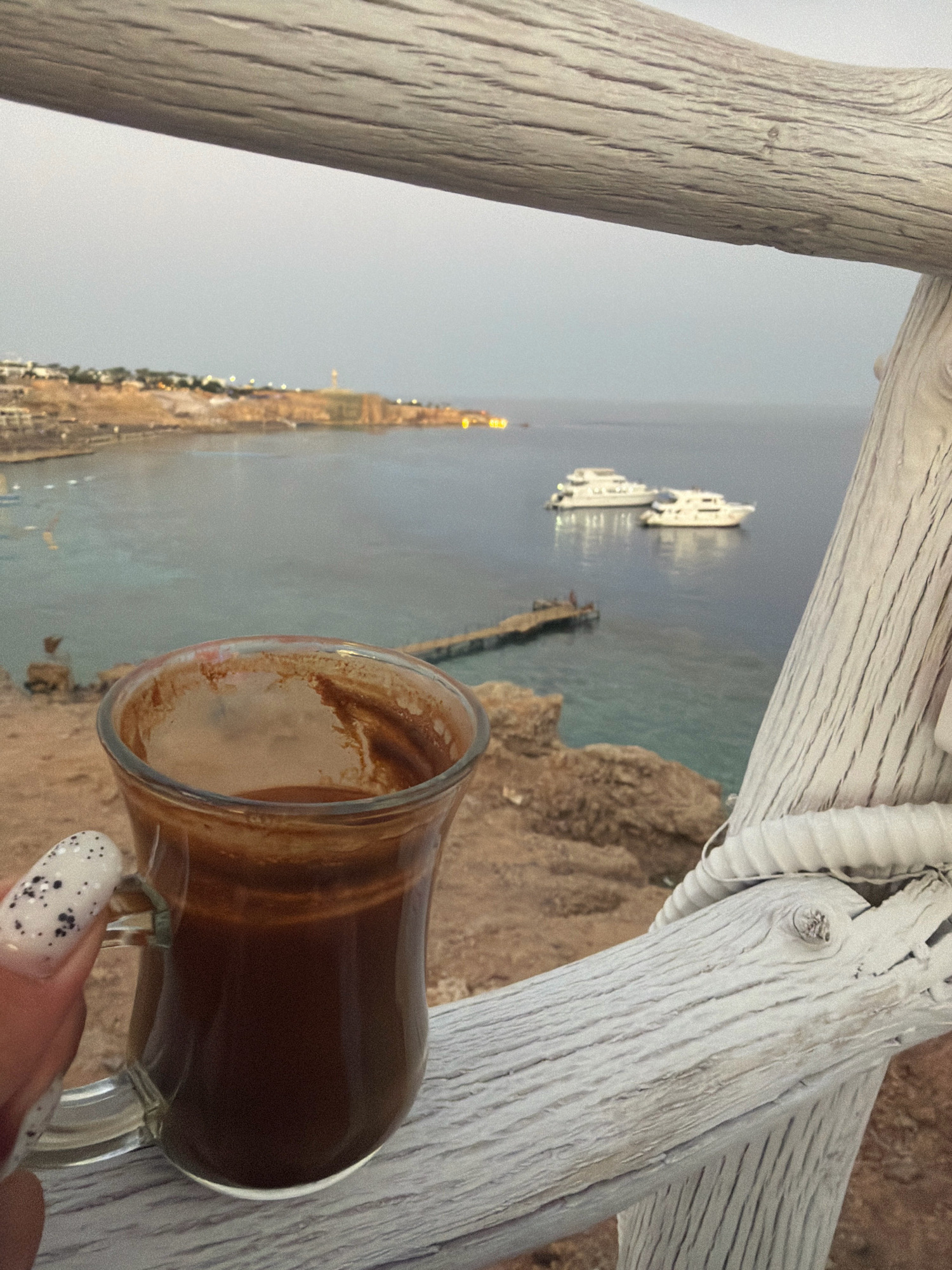 A close-up shot captures a clear glass mug of dark, frothy coffee held by a hand with white nail polish decorated with black speckles. The mug is positioned in the foreground, resting on a rustic, weathered wooden railing painted white. Beyond the railing, a scenic view unfolds of the Red Sea coastline in Sharm El Sheikh, Egypt. The water is a tranquil blue, dotted with two white yachts anchored near a small wooden pier. The distant shore is lined with buildings and illuminated lights, suggesting a resort town. The sky is overcast and a soft, diffused light suggests either dawn or dusk. The overall scene evokes a sense of peaceful relaxation by the sea.