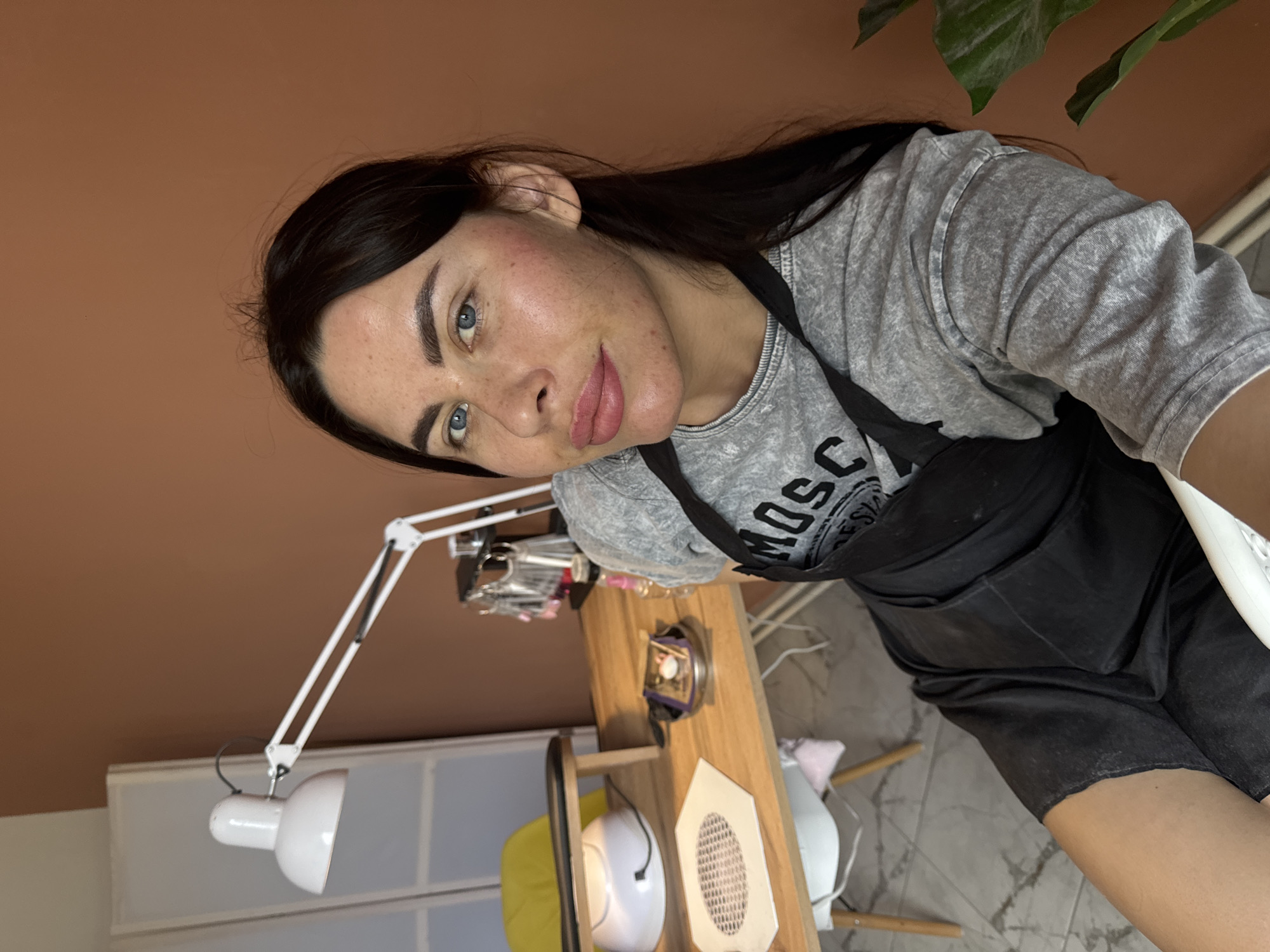 A medium shot captures a woman with dark hair and blue eyes sitting in what appears to be a nail salon. She is wearing a grey t-shirt with the word "MOSCHINO" printed on it and a black apron. The woman is looking at the camera with a neutral expression. Behind her, a wooden table is set up with various nail care tools, including a UV lamp, nail files, and bottles of polish. A white desk lamp is positioned above the table. The background features a brown wall and a tall, white screen or divider. A large green plant is visible on the right side of the frame. The scene suggests a professional setting, likely a beauty salon, and the overall ambiance is clean and organized. The location is identified as Chaikovsky, Russia.
