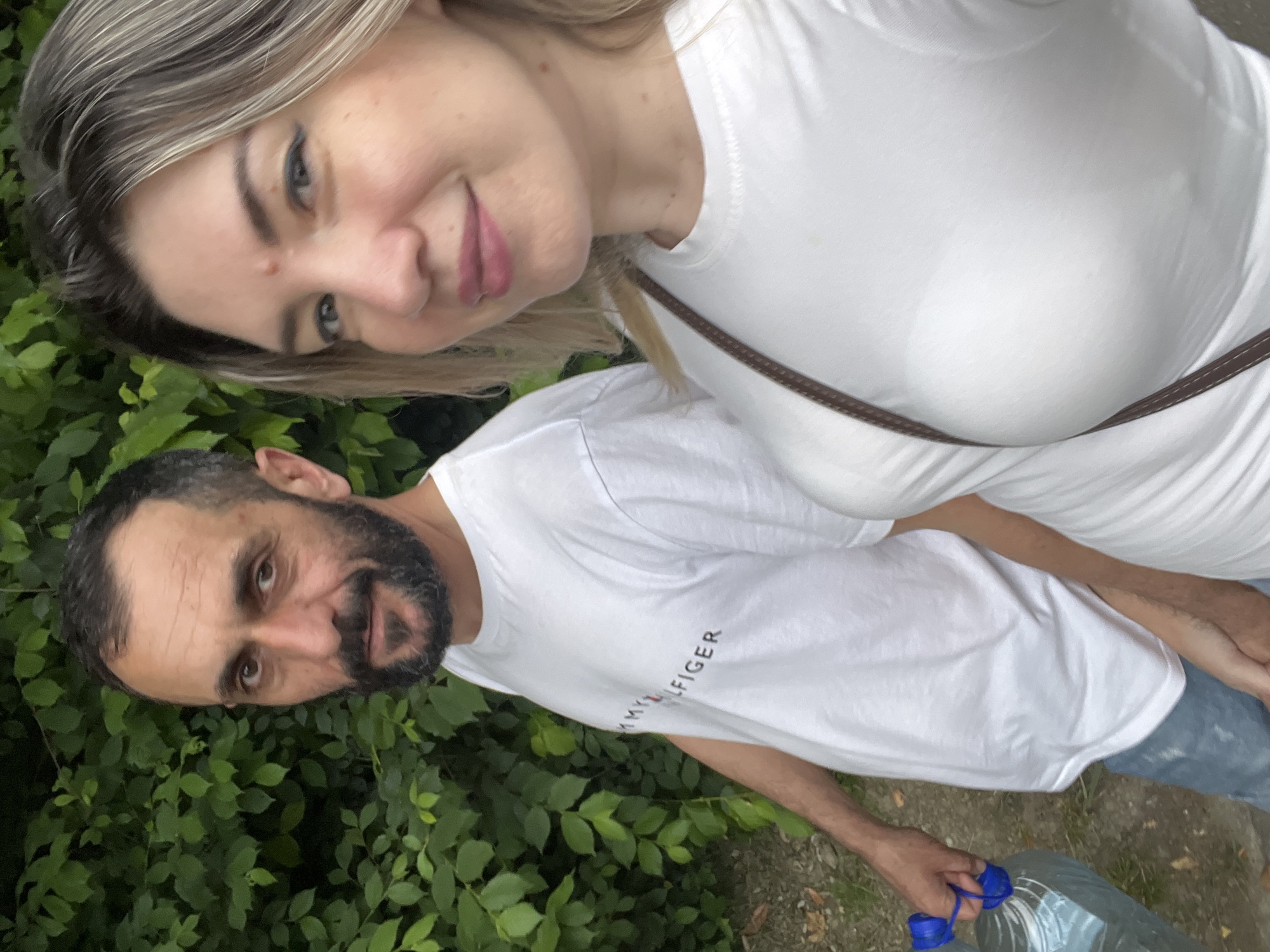 This media file shows two people, a woman and a man, in what appears to be an outdoor setting surrounded by lush green foliage. The woman, with blonde hair and fair skin, is positioned in the foreground, leaning into the frame. She is wearing a white t-shirt and has a gentle smile. Her hair is styled loosely, framing her face.
The man, with dark hair and a beard, is to her side, also looking towards the camera. He has a beard and appears to be wearing a white t-shirt with the text "TOMMY HILFIGER" visible across it. He is holding a large plastic bottle of water with a blue handle.
The scene is well-lit, suggesting it is daytime, and the foliage indicates an outdoor location, possibly a park or a wooded area. The overall mood appears relaxed and informal, possibly indicating a casual outing or a moment captured during an activity. The people are close to each other, suggesting a comfortable relationship. The focus is on the faces of the individuals and the immediate surroundings, with the background softly blurred.