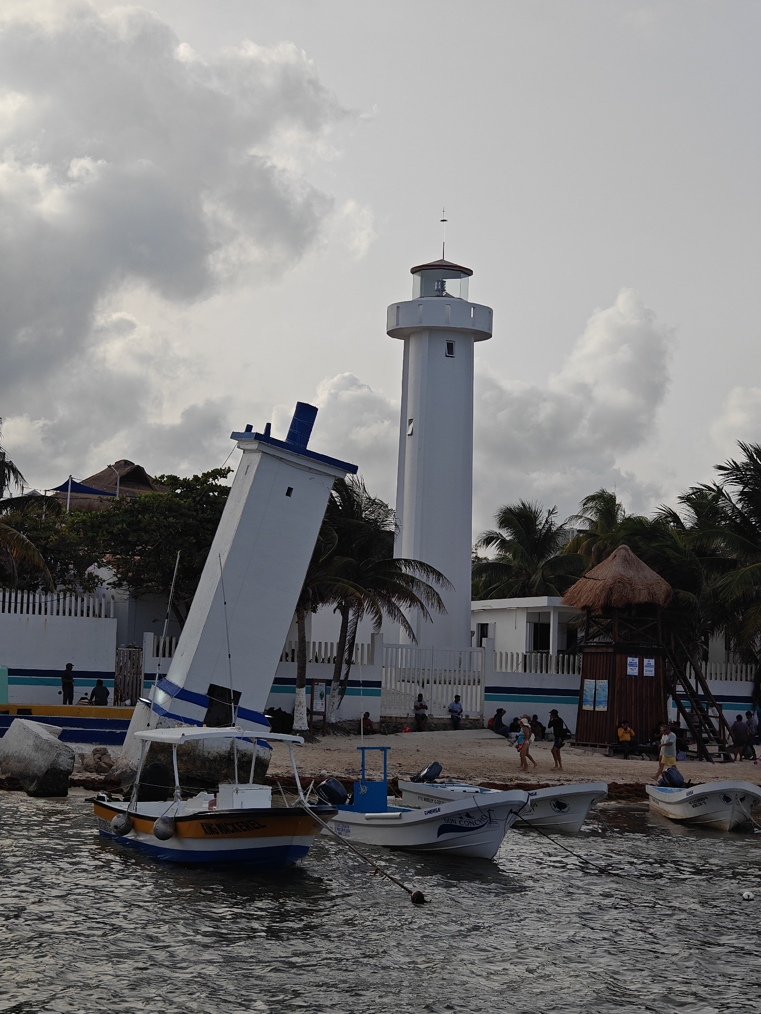 In Puerto Morelos, Mexico, a tilted lighthouse leans precariously on a sandy beach near the water's edge. A second, upright lighthouse stands tall in the background, a stark contrast to its fallen counterpart. Several small boats are moored in the shallow water, their reflections shimmering on the surface. Palm trees sway gently in the breeze, providing a tropical backdrop to the scene. A few people are visible on the beach, some relaxing, others walking along the shore. The sky is overcast with dramatic clouds, suggesting a change in weather is approaching. The overall impression is one of a picturesque coastal town, with a touch of dramatic visual interest due to the unusual state of the lighthouse.