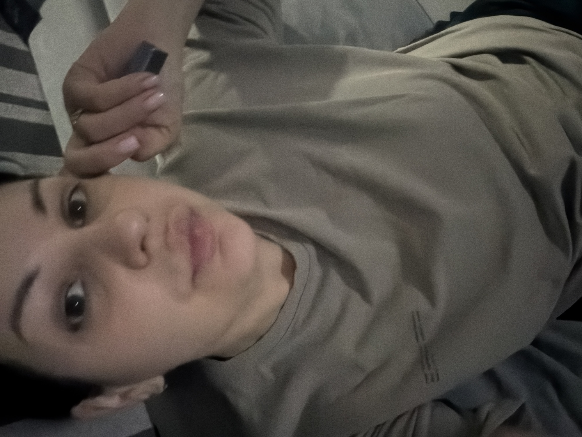 A woman is shown in a selfie, with her right hand touching her forehead. She is wearing a muted tan t-shirt. A small dark rectangular object is held between her thumb and forefinger. The background is blurred, with hints of striped upholstery suggesting a domestic setting, possibly a sofa. The lighting is dim, creating a grainy effect in the image. The woman's facial expression is neutral. The location is not discernible from the image.