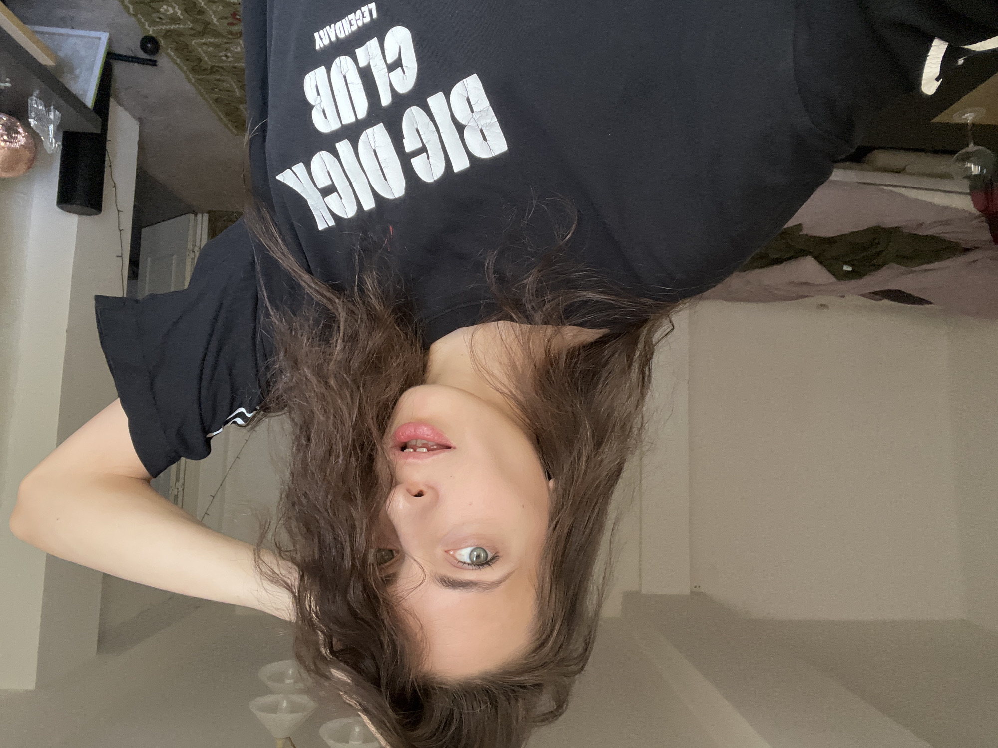 A woman with brown, wavy hair is seen in a selfie taken upside down. She wears a black t-shirt with the words "BIG DICK CLUB LEGENDARY" printed in white. Her eyes are light-colored, and she is slightly smiling. She is indoors with white walls. In the background, there is furniture and items on shelves. The location is Unknown, Unknown.