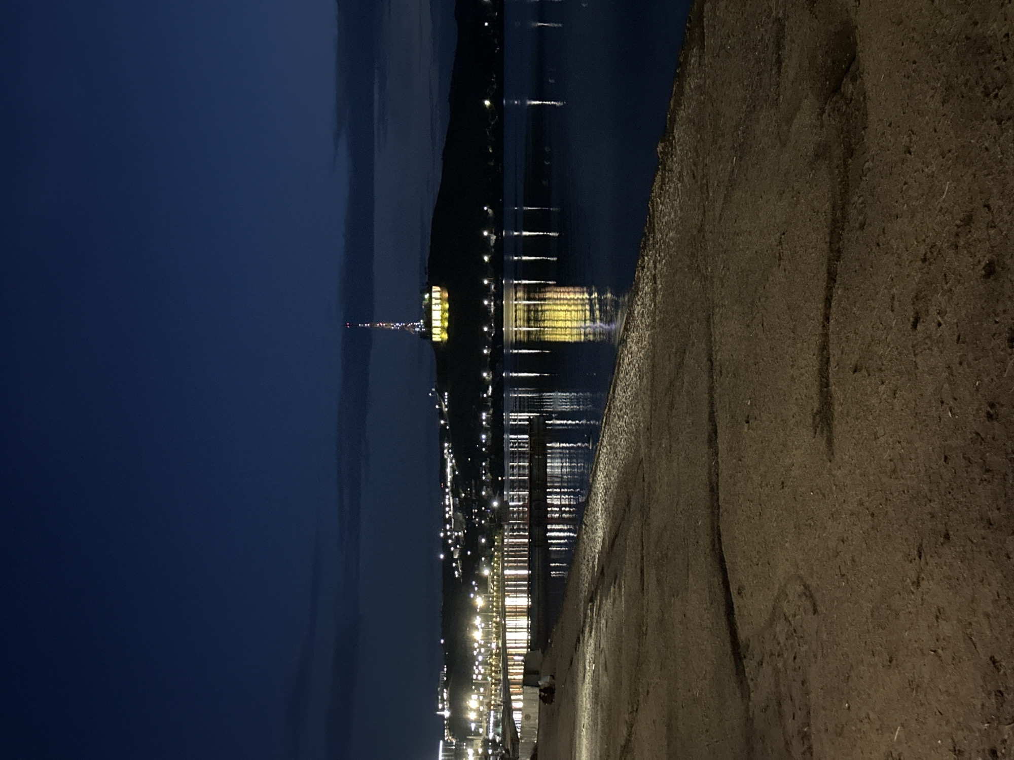 The image depicts a nighttime scene at Kokshetaū Bay in Kokshetau, Kazakhstan. In the foreground, a textured, sandy embankment occupies the bottom half of the frame. Beyond this, a body of water reflects the lights of a city skyline that stretches across a dark, hilly landscape in the background. The city lights are numerous and appear as bright, white and yellow points scattered along the elevated terrain. Dominating the skyline is a tall tower, illuminated with colored lights, and a large sign with illuminated yellow lettering reading "KOKSHETAU" is visible at its base. The water's surface is calm, mirroring the lights from the city and the tower. In the mid-ground, on the embankment near the water's edge, two figures are seated, silhouetted against the illuminated waterfront. The sky above is a deep, dark blue, with faint cloud formations visible on the horizon.