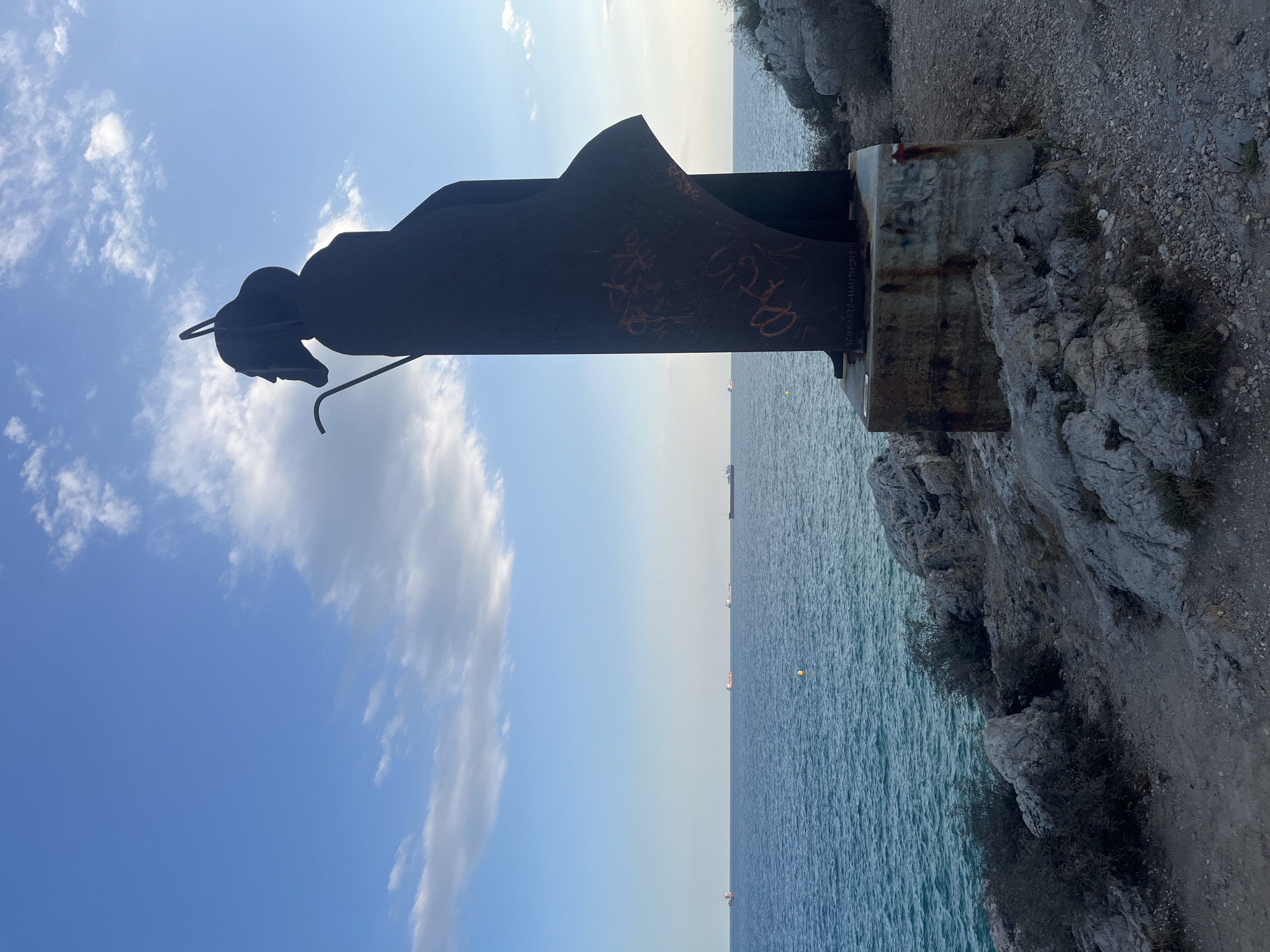 A dark metal sculpture of a robed figure, possibly a saint or shepherd, stands on a rocky cliff overlooking the Mediterranean Sea. The figure has a halo-like element above its head and is holding a staff. The sculpture is weathered and tagged with graffiti. In the distance, several cargo ships are visible on the water. The sky is blue with scattered white clouds. The scene is located at the Mirador del Palau in El Mirador, Spain.