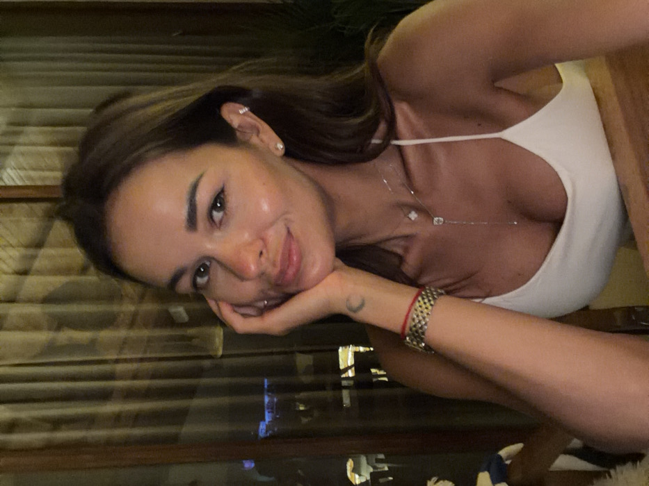 A woman leans on her hand, looking directly at the camera with a slight smile. She wears a white top with thin straps and a delicate necklace. A gold watch and red string bracelet adorn her wrist, complemented by a crescent moon tattoo. Her hair is brown with lighter highlights, framing her face. Background is blurred, featuring hints of blinds or window slats and city lights at night.