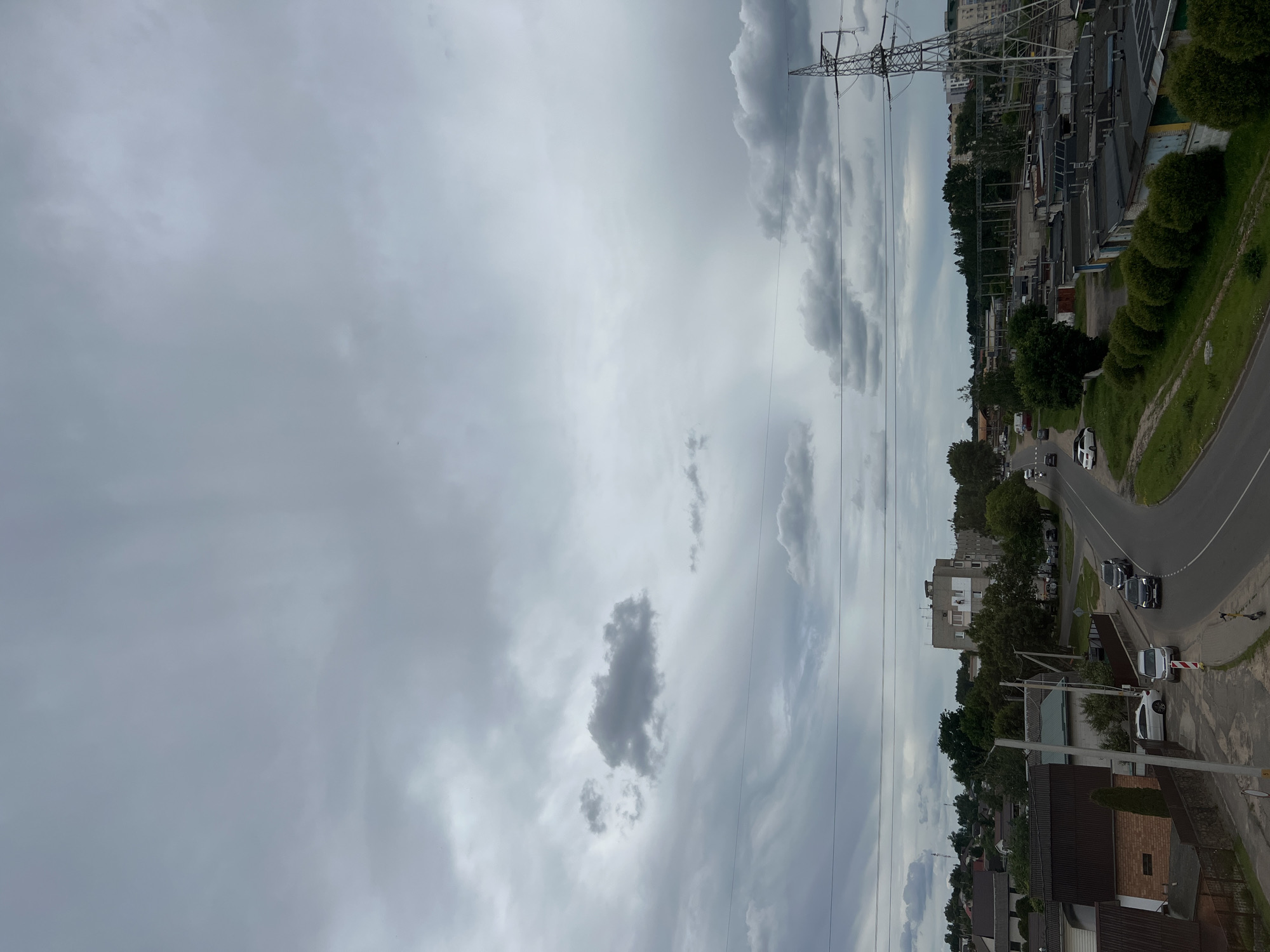 This media file captures a view of an urban or semi-urban landscape in Ждановіцкі сельскі Савет, Belarus, under an overcast sky. The scene is dominated by a dramatic sky filled with dark, heavy clouds, suggesting impending rain or a recent downpour. A prominent electricity pylon stands tall on the right side of the frame, with power lines stretching across the sky.
Below the sky, a road curves through the landscape. Several cars are visible on the road, indicating moderate traffic. To the right of the road, there are buildings, including what appear to be garages or small industrial structures with corrugated roofs, interspersed with some greenery and manicured bushes. Further back, apartment buildings and trees suggest a populated area. The overall impression is of a slightly gloomy, perhaps late afternoon or early evening, as indicated by the diffused light and heavy cloud cover. The setting appears to be a residential or mixed-use area on the outskirts of a town or city. There are no discernible people or specific events taking place, beyond the general activity of the road and the static presence of the infrastructure and buildings.