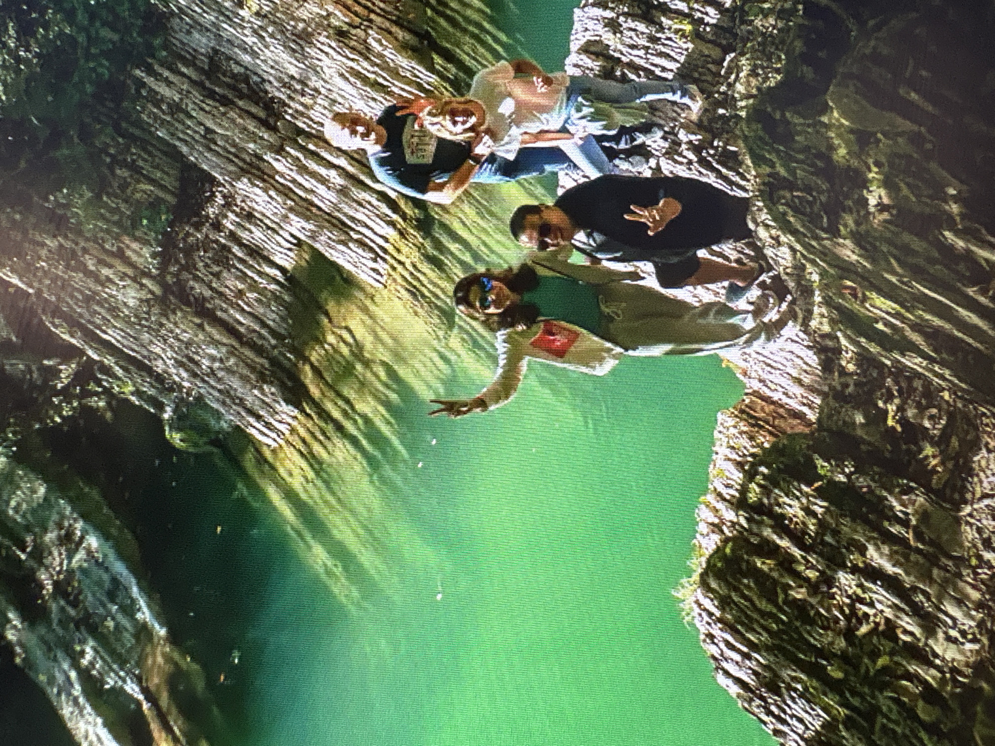 The image depicts four individuals posing for a photograph on a rocky outcrop overlooking a vibrant green body of water. In the foreground, two individuals are more prominently visible. The person on the left, wearing sunglasses and a light-colored jacket over a turquoise top, is gesturing with their left hand raised, fingers spread. To their right and slightly behind, a person in a dark jacket is also making a gesture with their hand, showing three fingers.
Behind these two, two more individuals are standing. A person in a dark blue t-shirt and blue jeans stands on the right, with their arm around the shoulders of a person in a light pink t-shirt and blue jeans. The person in the pink t-shirt is also making a "peace" sign with their hand.
The environment is characterized by rugged, textured rock formations that appear to be layered and jagged. The water is a striking emerald green, with dappled sunlight creating patterns on its surface. The background is filled with foliage and more rock faces, suggesting a natural, possibly mountainous or canyon-like setting.
There is no indication that this scene is located in the city of Moscow, Russia.