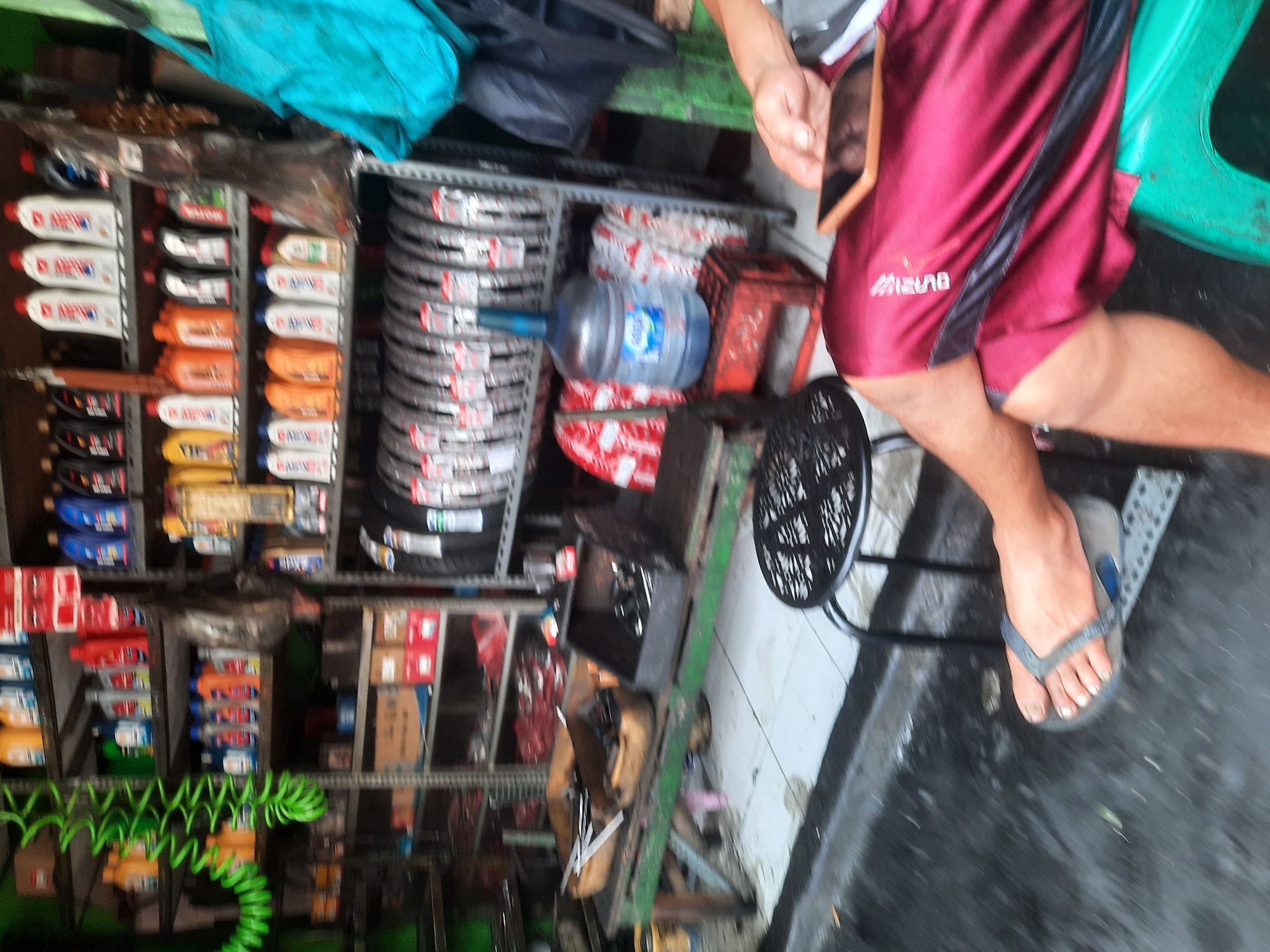 The image depicts an outdoor scene, likely a street-side stall in North Jakarta, Indonesia. The primary focus is on a selection of automotive fluids and tires displayed on shelving units. Several bottles of various colored liquids, presumably motor oil or additives, are stacked on shelves. A large water jug is also visible, placed on a red crate. In the foreground, a person's legs and feet are visible, with one foot wearing a flip-flop. The individual appears to be sitting or resting, with a mobile phone placed on their lap, resting on a maroon garment. The environment suggests a commercial area, with some elements of disarray, such as scattered items on a wooden table and a wet ground surface, indicating recent rain or cleaning. The overall atmosphere is that of a busy, practical marketplace catering to automotive needs.