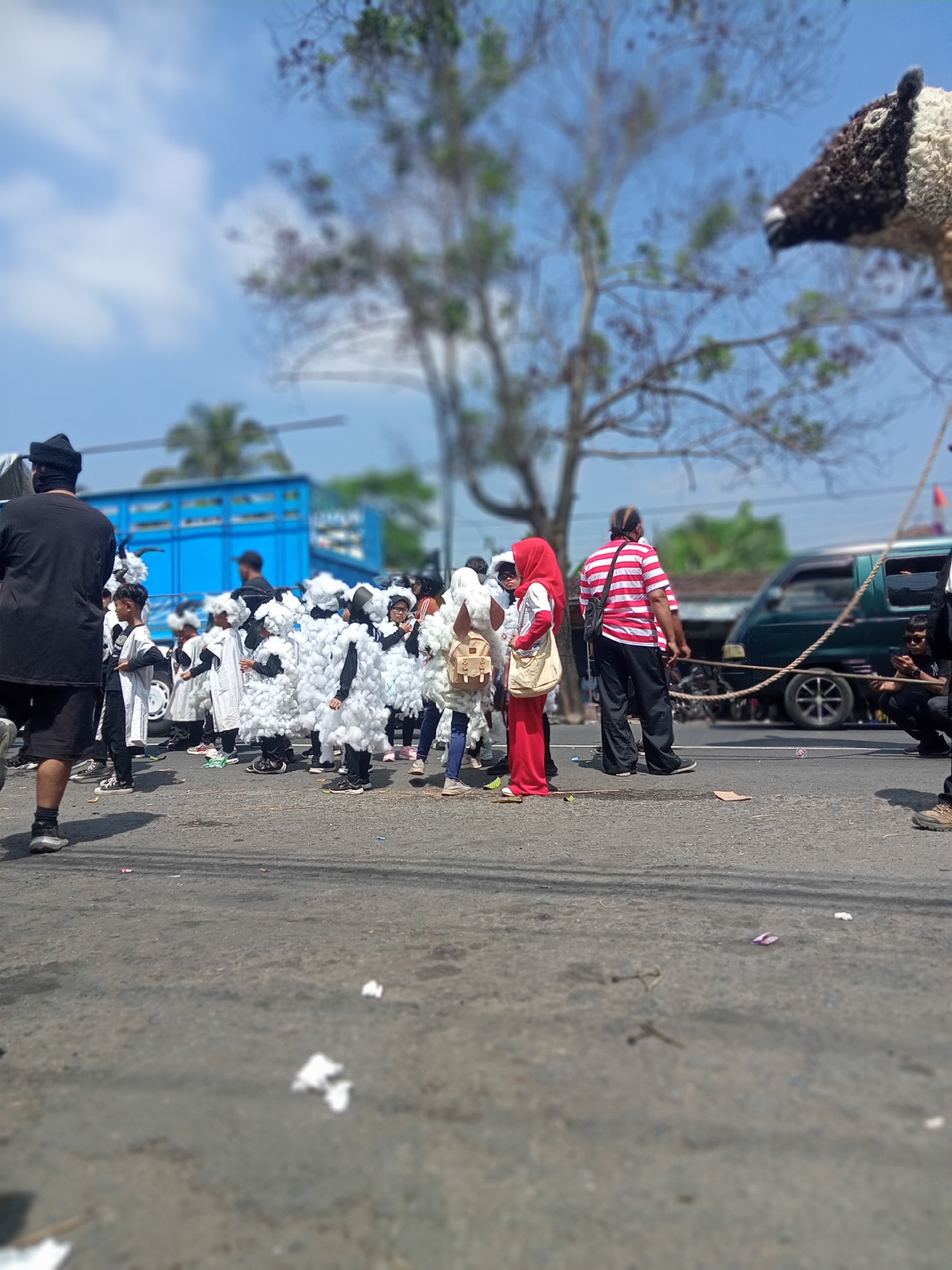 In Urutsewu, Indonesia, a street parade is underway, featuring a group of children dressed as sheep. The children are wearing white, fluffy costumes that resemble sheep's wool, complete with headpieces. They are standing in formation on an asphalt road, some holding onto each other. Behind them, adults are observing the procession. One adult in the foreground, viewed from the back, is wearing a black head covering and black clothing. Another adult in the background wears a red and white striped shirt and black pants. A large blue truck is parked on the left side of the road, and a dark green van is visible further down the street on the right. The sky is blue with scattered white clouds, and trees with sparse foliage line the background. The ground is littered with small pieces of white debris. The scene suggests a cultural event or celebration taking place in a semi-urban or rural setting.