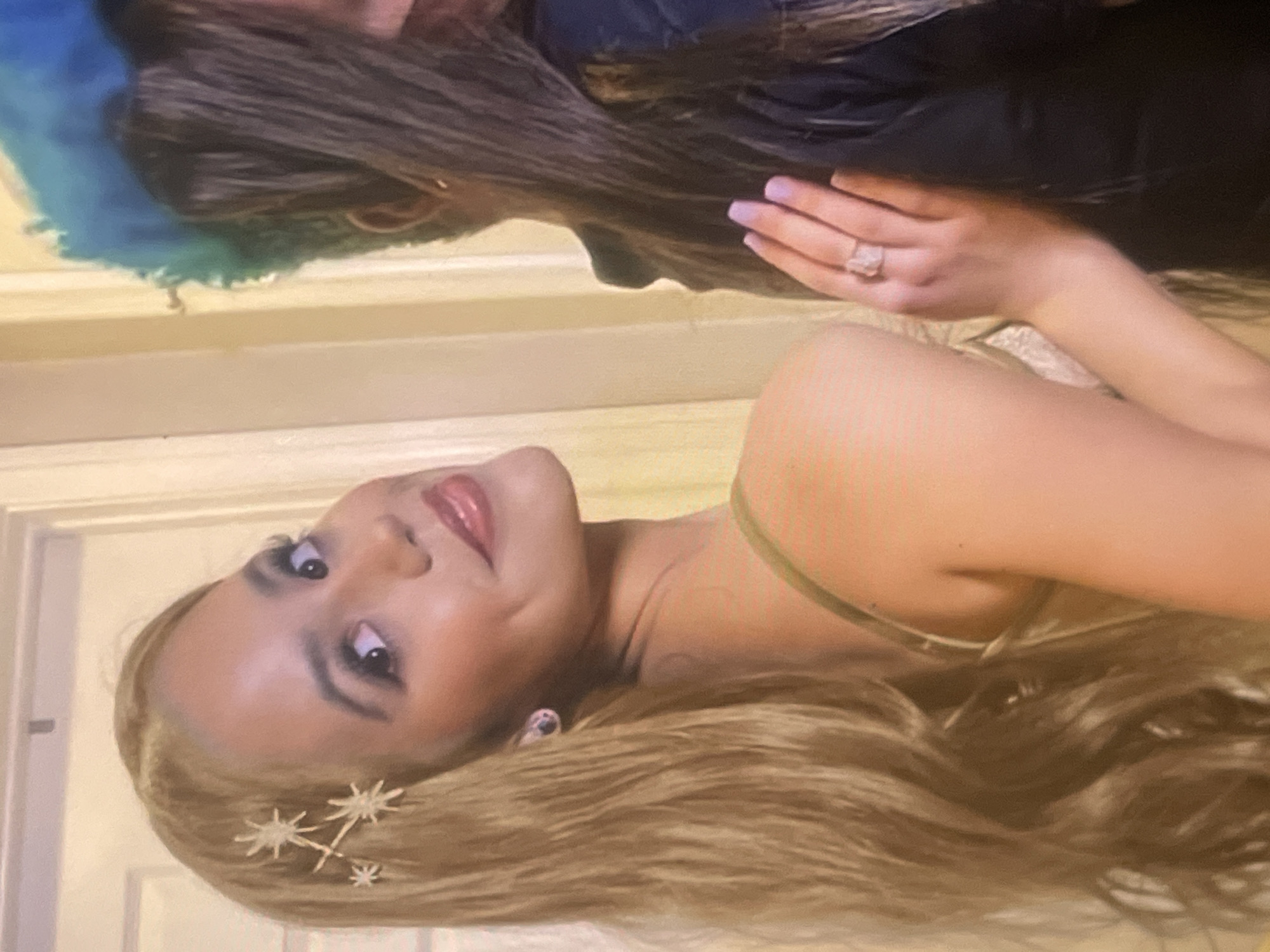 The image displays a close-up portrait of a young woman with long, wavy blonde hair, adorned with several star-shaped hair accessories near her temple. She looks slightly upwards and to her left, offering a subtle, gentle smile. Her makeup includes dark eyeliner, eyeshadow, and glossy pinkish-red lipstick. She wears a thin-strapped, light-colored top, possibly in a champagne or gold hue.
In the upper background, a reflection reveals another individual. This person has dark brown hair and is dressed in a dark garment. Their left hand is visible, featuring a prominent, sparkling ring on the ring finger and nails painted a light lavender color. A vibrant blue and green, possibly fluffy or feathery, object or fabric is also visible in the reflection next to the person's head.
The setting appears to be indoors, against a light-colored wall with white architectural trim, suggesting a room or event space. The overall impression is a posed photograph, possibly a selfie, with the reflected person potentially holding the camera. The scene is situated in Poltava, Ukraine. No text is visible in the image.