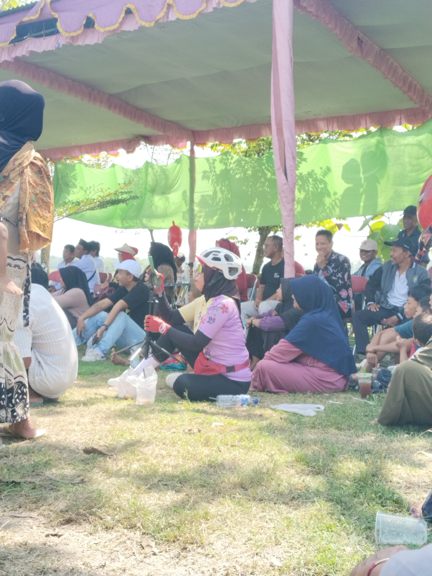 The image shows a gathering of people outdoors under a tent-like structure with pink and green decorative trim. The location is Karanganom, Indonesia.
People of varying ages and genders are seated on the grass and on chairs, some are wearing head coverings and helmets. They appear to be observing something.
Objects present include water bottles, drinks in plastic cups, and possibly camera equipment. The environment is grassy with trees visible in the background.