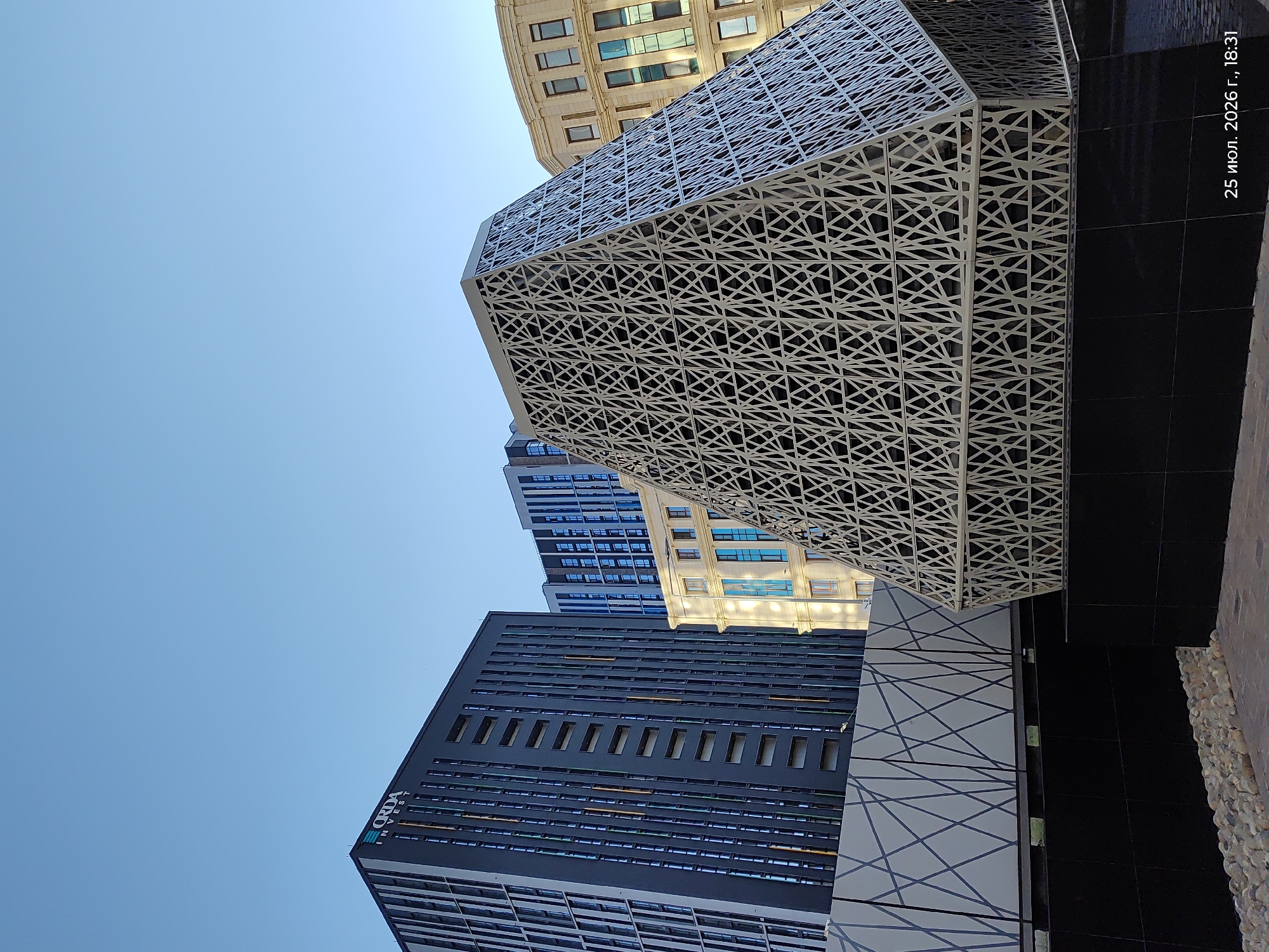 The image captures a modern architectural scene in Astana, Kazakhstan, under a clear blue sky. Dominating the foreground is a uniquely shaped, angular structure with a geometric, latticed white facade. Behind this, to the left, stands a tall, dark-colored skyscraper with vertical window lines and the "CRDA INVEST" logo visible near its top. To the right of the dark skyscraper, a lighter-colored, more classical building with arched windows is partially visible. Further back, another modern building with blue-tinted windows can be seen. The ground level features dark tiled surfaces and a section with decorative stones. A timestamp in the lower right corner reads "25 июл. 2026 г., 18:31", indicating the date and time the photograph was taken.