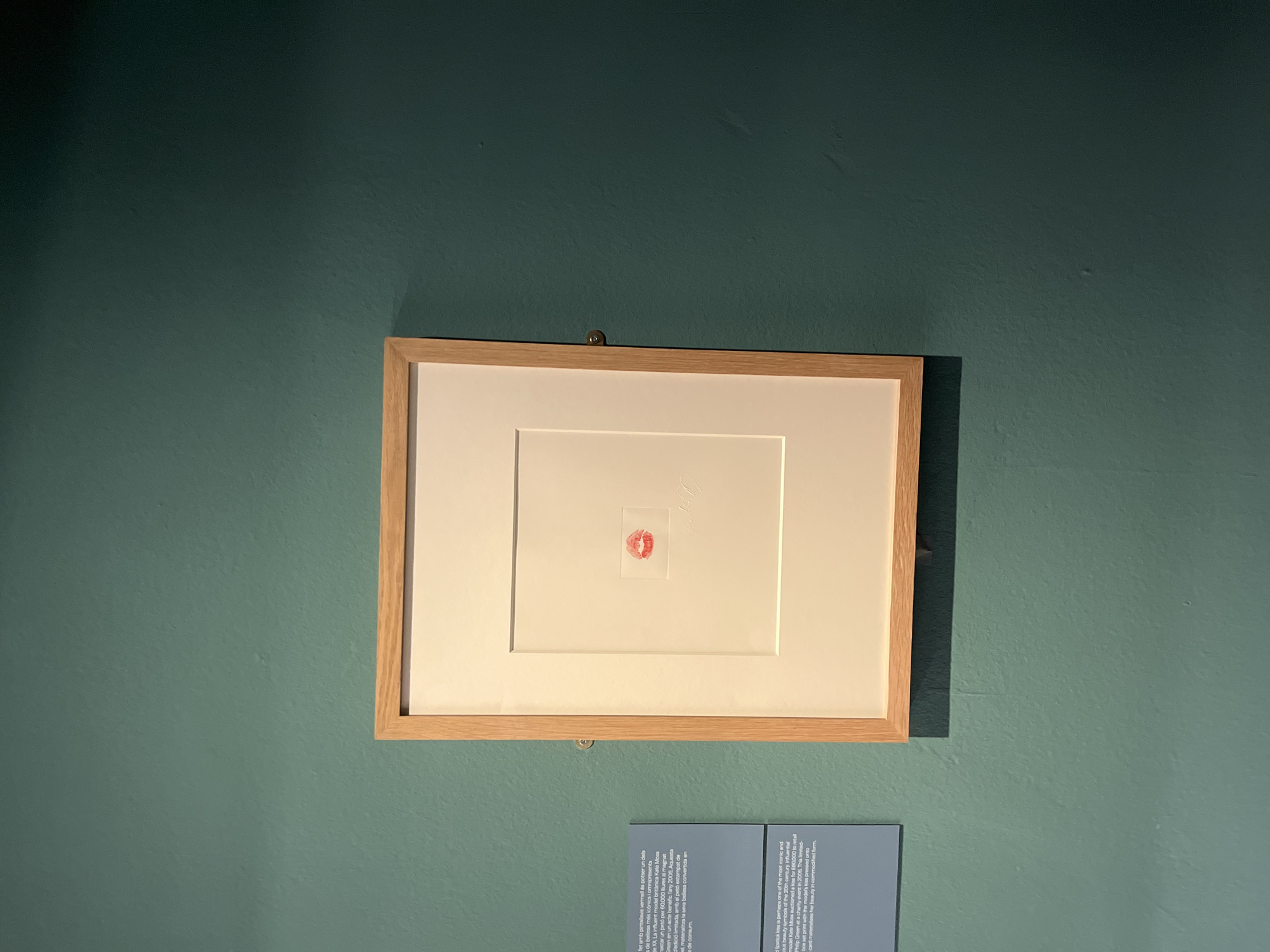 The image depicts a framed artwork displayed on a teal-colored wall in a museum or gallery. The artwork itself is a minimalist print featuring a red lipstick kiss mark. The print is matted with a thick, cream-colored border and encased in a light wood frame. To the left of the framed artwork, a small informational plaque with text is visible. This scene is located in Barcelona, Spain, specifically within the Picasso Museum, as indicated by the informational plaque. The museum is renowned for its extensive collection of works by the Spanish artist Pablo Picasso. The lighting in the scene is subdued, casting a shadow from the framed artwork onto the wall. The overall impression is one of quiet contemplation and appreciation for the art on display.