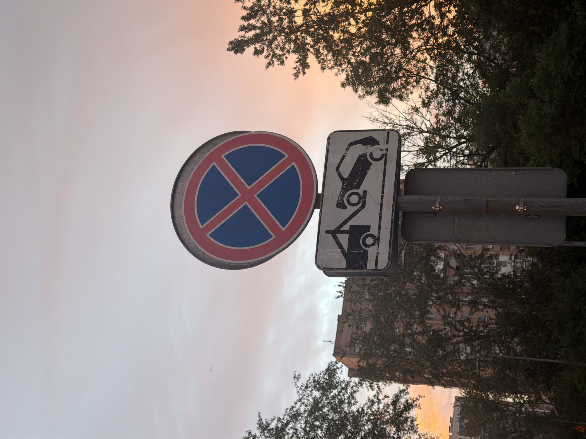 A low-angle, full shot captures a street sign against a pale pink and orange sunset sky. The sign, mounted on a metal pole, consists of two rectangular panels. The top panel is a circular red border with a blue interior, crossed by two red diagonal lines, indicating "no parking." Below it, a rectangular white panel displays a black silhouette of a tow truck hoisting a car, signifying that vehicles parked here will be towed. Trees with dark green foliage frame the sign, and a tall, multi-story brick building is visible behind the left side of the sign. The scene is set in a city environment, with a clear indication of an urban area.