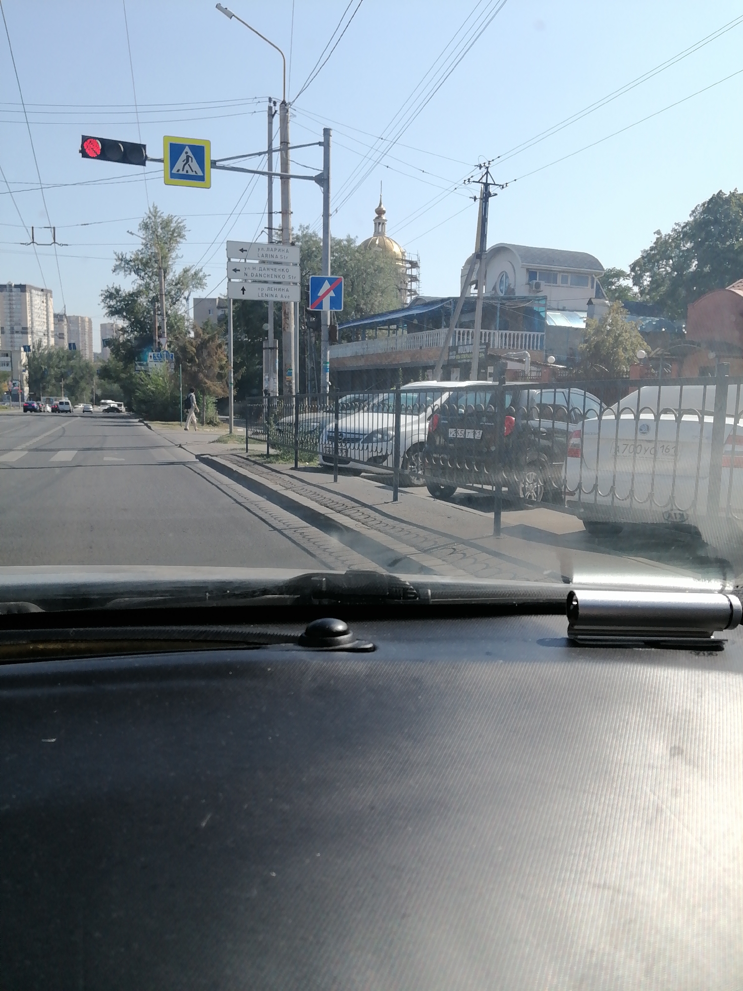 The image was taken on a sunny day at an intersection in Rostov-on-Don, Russia. The view is from inside a vehicle, looking forward through the windshield and dashboard. In the foreground, the textured black dashboard and a silver clip are visible.
The middle ground shows a street with cars and pedestrian crossings. A traffic light is visible with a red light illuminated. A pedestrian crossing sign with a blue square and a white silhouette of a person walking is also present. Several street signs are mounted on a pole. One sign indicates "ул. ЛАРИНА LARINA Str." with an arrow pointing left. Another sign indicates "ул. Н. ДАНЧЕНКО N. DANCHENKO Str." with an arrow pointing left. A third sign indicates "пр. ЛЕНИНА LENINA Ave." with an arrow pointing up. A blue sign with a white arrow and a red diagonal line, indicating a no-entry zone, is also visible.
Further down the street, a black car and a white car are parked behind a black metal fence. In the background, apartment buildings and trees line the street. A golden dome, likely from a church or a historical building, is visible above the trees. The sky is clear and blue.