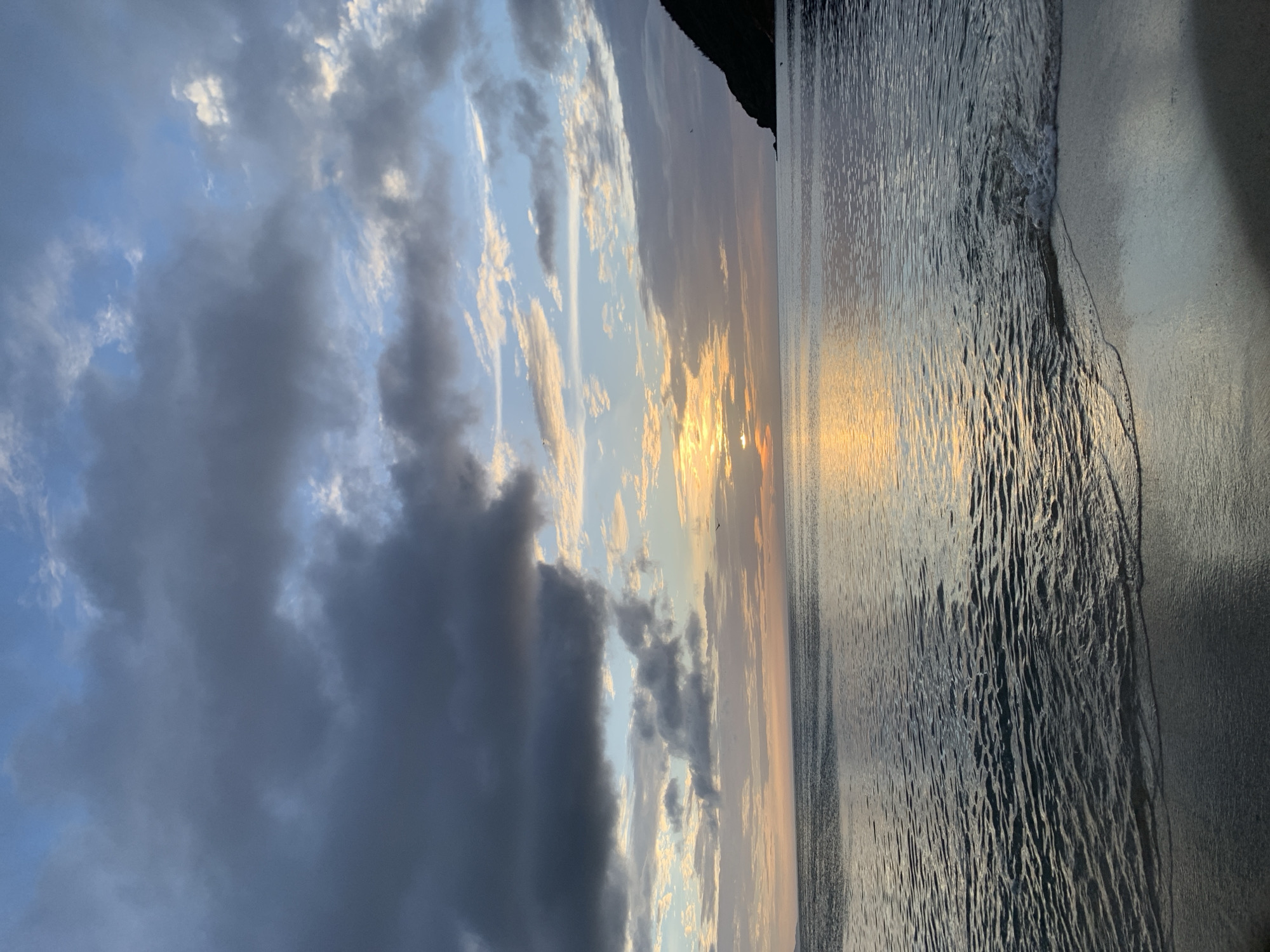 A serene view of the ocean at dusk, with waves gently lapping at a sandy shore. The sky is a mix of dark, dramatic clouds and softer, lighter ones, with the setting sun casting a warm, golden glow on the horizon. A silhouette of a lush green hill can be seen on the right side of the frame, bordering the water. The scene captures the tranquil beauty of a coastal evening in Juan Griego, Venezuela.
