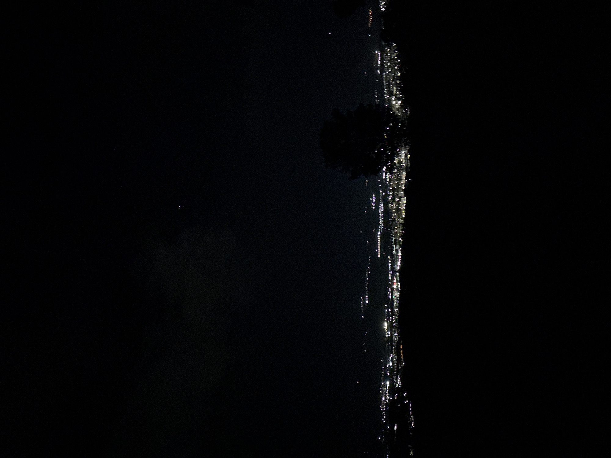 The image shows a dark night scene with a vertical strip of lights along the right side. The lights appear to be from a city, possibly reflected in water. There is a dark, blurry shape that could be a tree silhouette above the lights. The image is dark and lacks detail, making it difficult to discern specifics. The location is identified as Marulevo, Bulgaria, but there is no additional information about the environment.