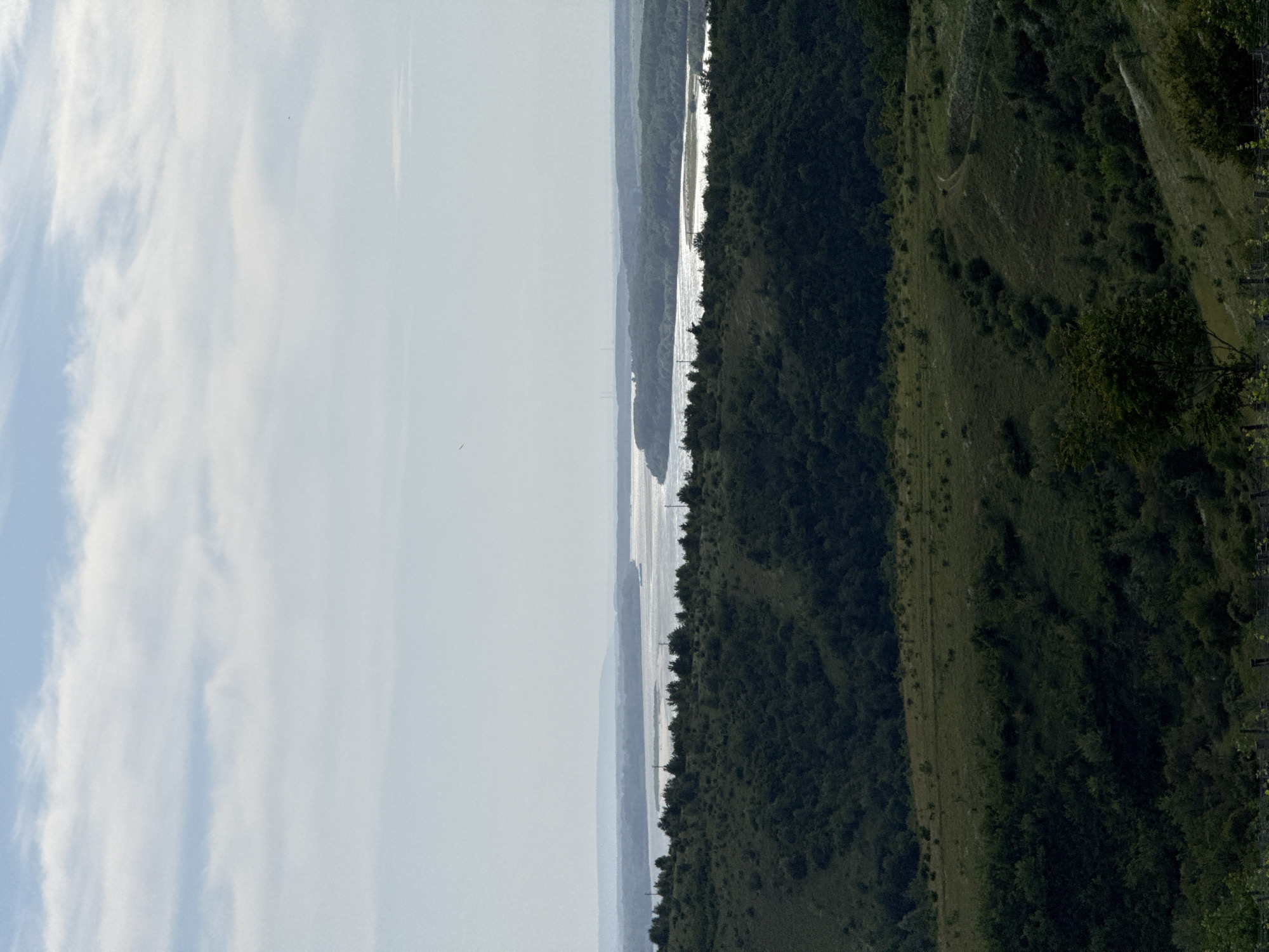 The image shows a landscape in Mechka, Bulgaria. A body of water, likely a river or lake, runs vertically down the center of the frame, reflecting sunlight. To the left is a bright sky with wispy clouds. To the right, a heavily wooded area covers a hillside. A thin, light-colored path or clearing cuts through the vegetation along the slope. The overall impression is a scenic view combining water, sky, and forested terrain.