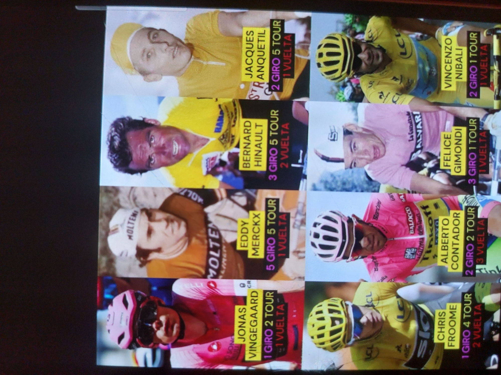 This media file captures a photo of a screen or display showcasing a grid of nine renowned professional cyclists. The image is rotated 90 degrees clockwise. The subjects are all male cyclists, each depicted in a headshot or upper body shot, wearing cycling jerseys and often helmets, representing different eras of the sport.
The scene is a collage celebrating cyclists who have achieved significant victories in Grand Tours. Each portrait is accompanied by the cyclist's name and a summary of their wins in the Giro d'Italia ("Giro"), Tour de France ("Tour"), and Vuelta a España ("Vuelta").
Notable details include:
- The top row features Jacques Anquetil (wearing a yellow jersey and cap) and Vincenzo Nibali (in a yellow jersey and helmet).
- The middle row shows Bernard Hinault (in a yellow jersey), Felice Gimondi (in a pink jersey and white cap), and Eddy Merckx (in an orange Molteni team jersey).
- The bottom row includes Alberto Contador (in a pink Tinkoff jersey and helmet), Jonas Vingegaard (in a pink jersey and helmet), and Chris Froome (in a yellow jersey and helmet).
- Cyclists are seen in varying attire, including yellow jerseys (Tour de France leader), pink jerseys (Giro d'Italia leader), and team jerseys.
- Most cyclists have neutral or focused expressions, typical of sports portraits.
Visible text associated with each cyclist and their Grand Tour victories:
- **JACQUES ANQUETIL:** 2 GIRO 5 TOUR 1 VUELTA
- **VINCENZO NIBALI:** 2 GIRO 1 TOUR 1 VUELTA
- **BERNARD HINAULT:** 3 GIRO 5 TOUR 2 VUELTA
- **FELICE GIMONDI:** 3 GIRO 1 TOUR 1 VUELTA
- **EDDY MERCKX:** 5 GIRO 5 TOUR 1 VUELTA
- **ALBERTO CONTADOR:** 2 GIRO 2 TOUR 3 VUELTA
- **JONAS VINGEGAARD:** 1 GIRO 2 TOUR 1 VUELTA
- **CHRIS FROOME:** 1 GIRO 4 TOUR 2 VUELTA
The image appears to be a personal photograph taken of a digital or printed display in an indoor setting, given the presence of glare and the screen-like texture.