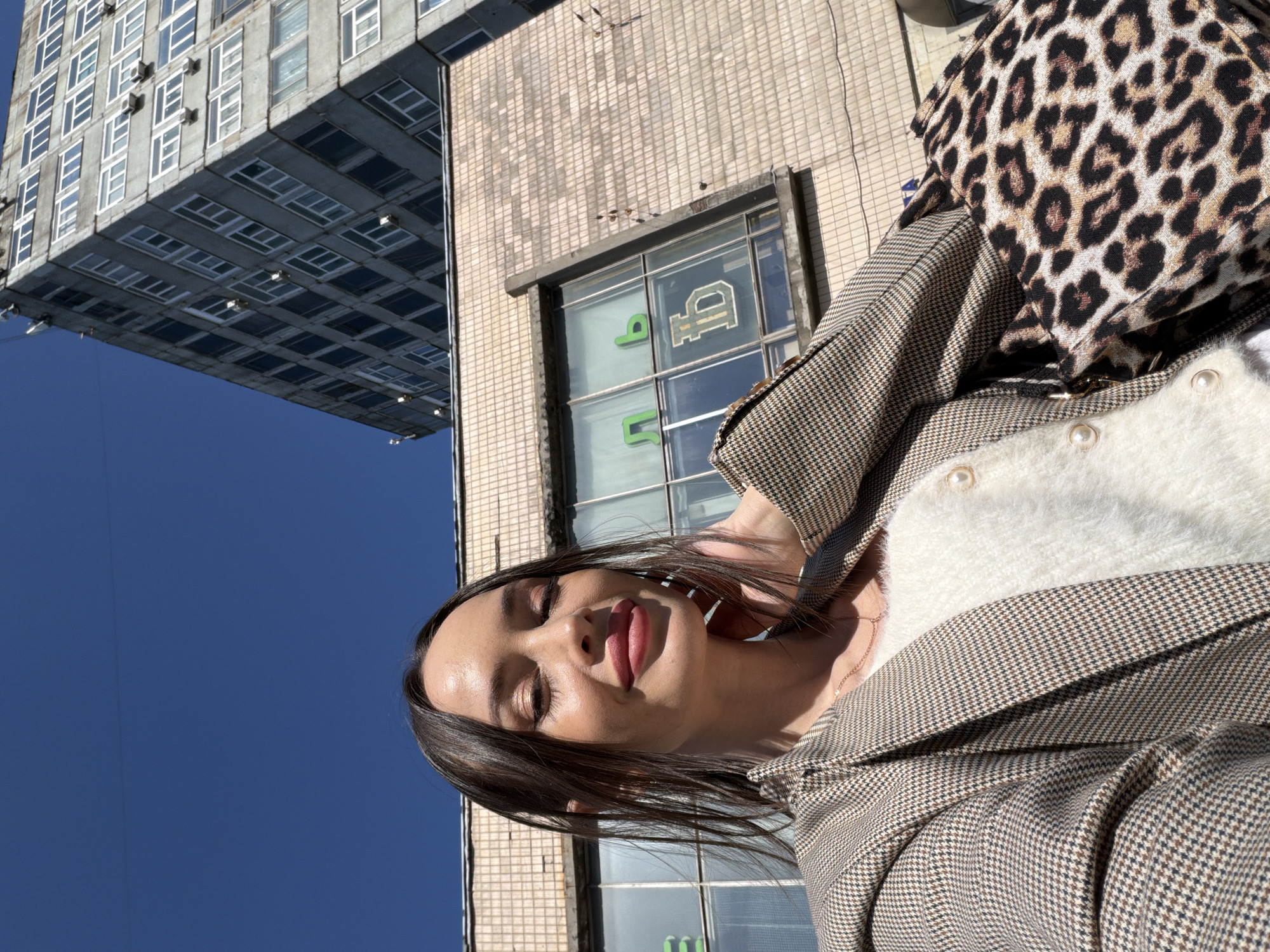 A young woman is taking a selfie. She wears a houndstooth blazer over a white sweater and carries a leopard-print bag. The background includes a building in Saint Petersburg, Russia, with Cyrillic script visible in the windows. The sky is clear and blue.