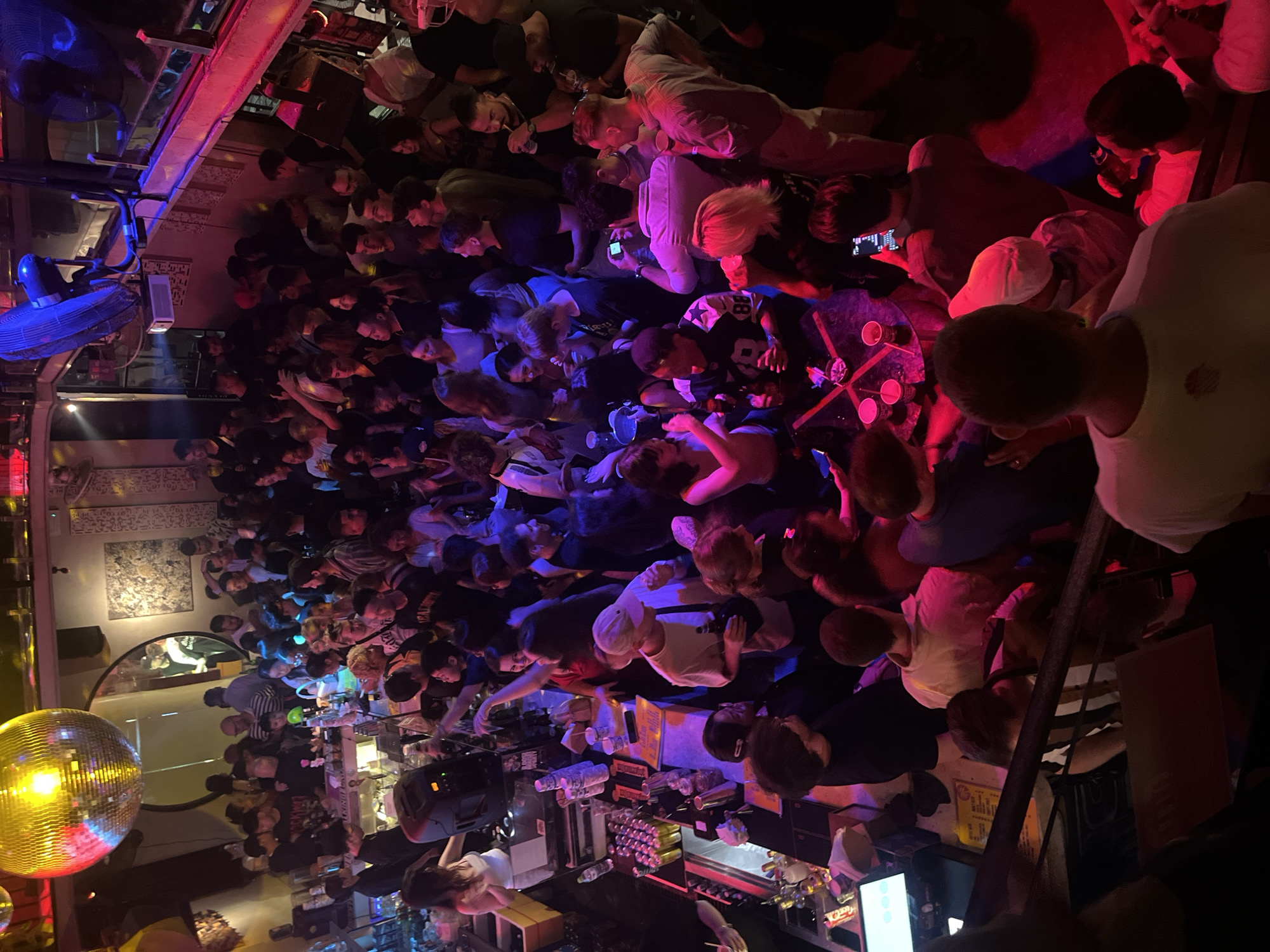 The image depicts a crowded indoor establishment, likely a bar or nightclub, with dim lighting and vibrant red and blue hues from stage lights. A large disco ball hangs from the ceiling, reflecting light throughout the space. Patrons, predominantly young adults, are gathered, socializing, dancing, and consuming beverages. Some individuals are at a bar area, where drinks are being served. The atmosphere appears lively and energetic. The scene is consistent with a nightlife venue in Đà Nẵng, Vietnam.