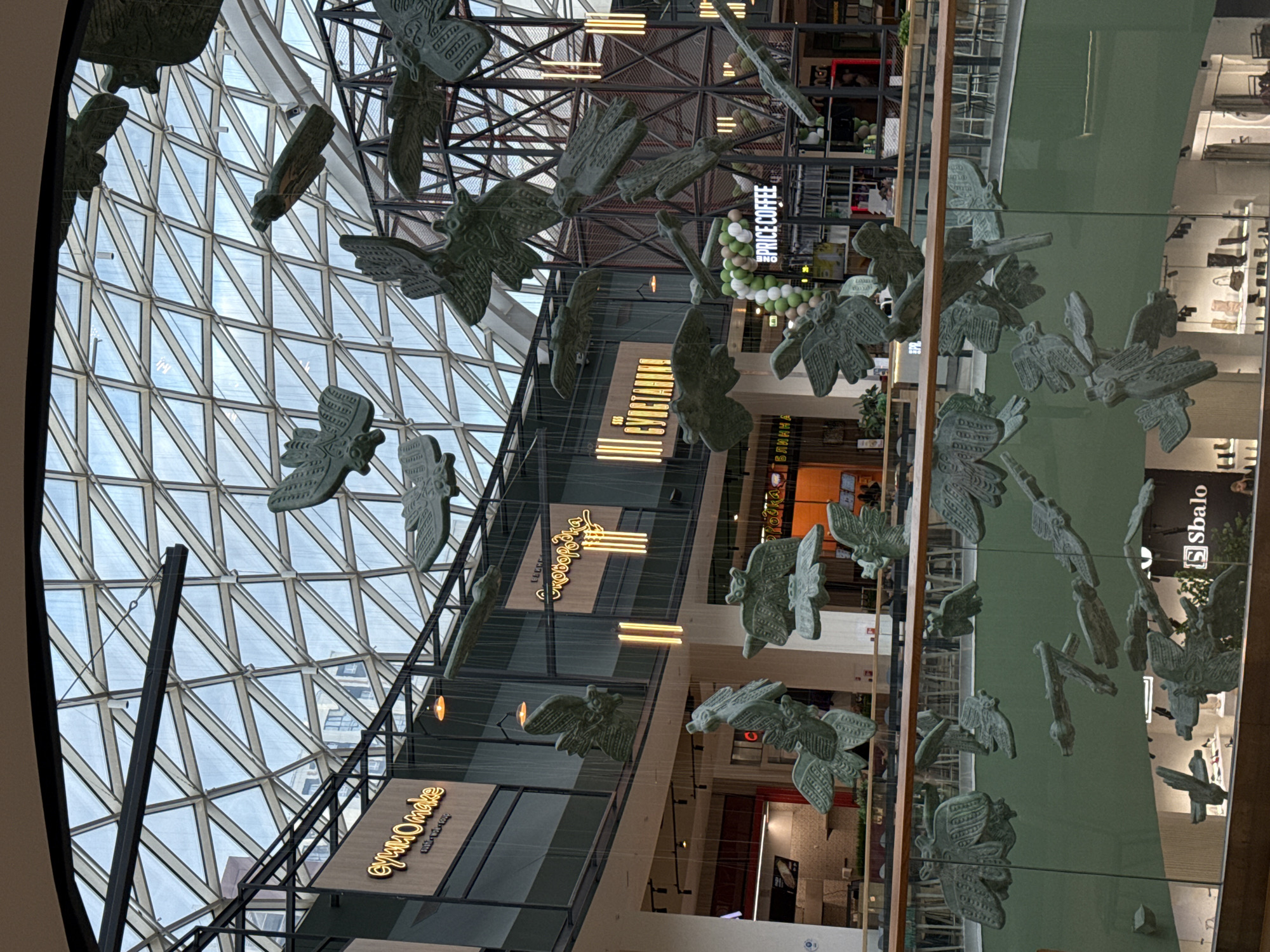 The image depicts the interior of a shopping mall with a high, triangular glass roof and metal support structures. Hanging from the ceiling are numerous sculpted bird-like figures. Storefronts are visible on multiple levels, including one for "PRICE COFFEE", "МимиОmake" and "Sbalo". The location is most likely in the city of Perm, Russia, based on the Cyrillic script used on store signs. No people are clearly visible in the image. The overall scene is a modern architectural design with an artistic installation.