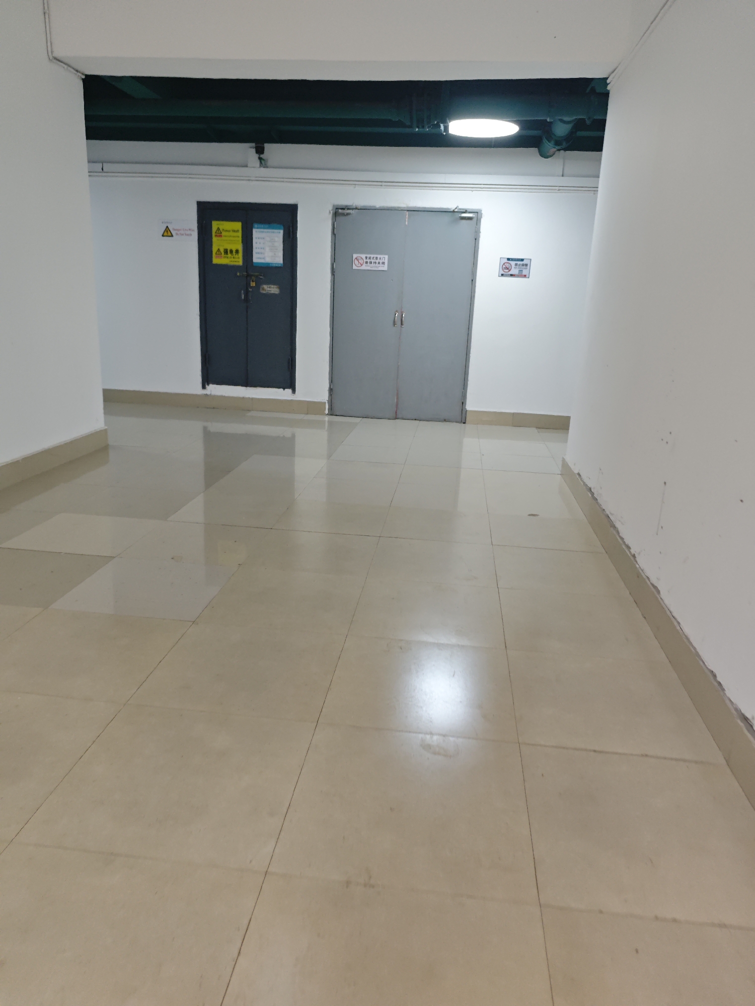 The image depicts an interior hallway in what appears to be a commercial or industrial building. The hallway is brightly lit with overhead fluorescent lights, one of which is visible towards the top of the frame. The walls are painted white and there is a darker band of what appears to be painted metal conduit running along the ceiling. The floor is tiled with large, light-colored square tiles, which are clean and reflect the light.
On the left side of the hallway, there are two dark gray doors side-by-side. The left door has a padlocked hasp and several warning signs and informational labels. One sign is a yellow triangle with a lightning bolt symbol. Another appears to be a safety instruction. The right door also has a padlocked hasp and a sign that reads in Chinese "严禁烟火" which translates to "No Smoking, No Open Flames".
On the right side of the hallway, further down, is a single, lighter gray door with two vertical handles. This door also has a sign with a red circle and a diagonal line over a cigarette, indicating "No Smoking", and Chinese text below.
The architecture suggests a functional space, possibly an electrical room or utility closet access. The overall environment is clean and well-maintained. The provided contextual information places this scene in the Chongqing High-tech Zone, China, implying it is likely part of a business or research facility within that zone. There are no people visible in the image.