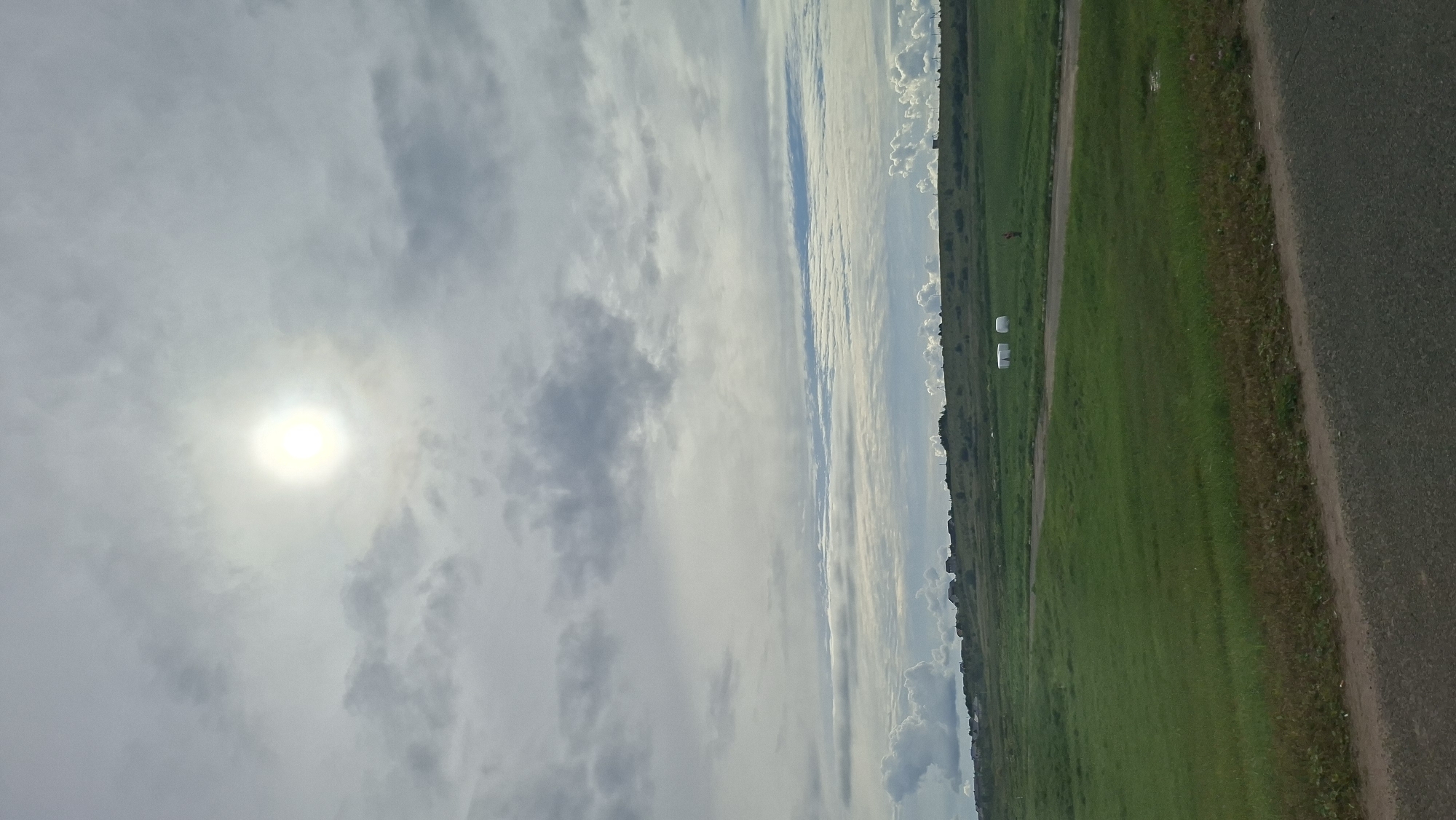 This is a vertical, eye-level, outdoor, full shot of a grassy landscape under a cloudy sky. The sun is a bright white disk in the upper middle of the frame, partially obscured by clouds. The sky is filled with a mix of light and dark gray clouds, with some lighter patches that suggest the sun's presence.
The foreground consists of a paved road with a gravel shoulder. Behind the road is a large expanse of green grass, which appears to be a field or a park. There are two white, cylindrical objects, possibly hay bales or storage units, located in the middle ground to the right. A small, red-orange object, possibly a person or marker, is situated between the two white objects. In the distance, a low line of hills is visible, with scattered houses and trees along the crest. The overall impression is a rural or semi-rural setting, likely in Myadzyel, Belarus, given the context. The lighting suggests it is daytime, but the cloudy conditions temper the brightness of the sun.