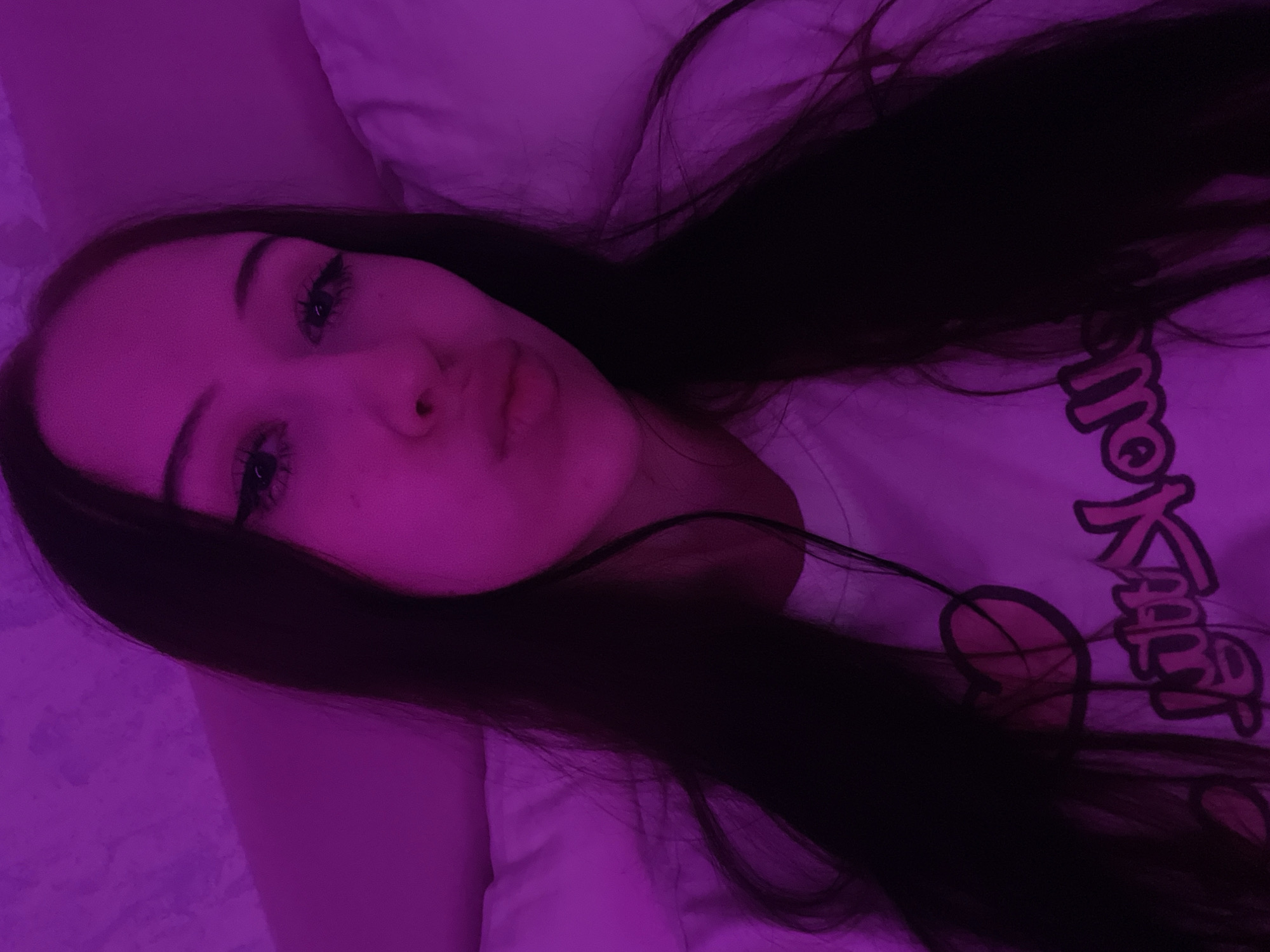The image shows a close-up, tilted selfie of a young woman lying down, with her face illuminated by purple light. She has dark hair that falls around her face and over her shoulder, partially obscuring a white t-shirt with pink text. The text appears to be in a stylized font, possibly Japanese characters, but it is not entirely clear due to the lighting and hair obstruction. The woman's eyes are open, and she is looking towards the camera. Her makeup is visible, including eyeliner and mascara. Her lips are slightly parted, and her expression could be interpreted as neutral or slightly playful. The background is a soft, indistinct texture, likely a pillow or bedding, also bathed in the purple light. The overall impression is of a casual, intimate moment, possibly taken at night or in a dimly lit room, given the dominant purple hue. No specific location cues are visible other than the general context provided.