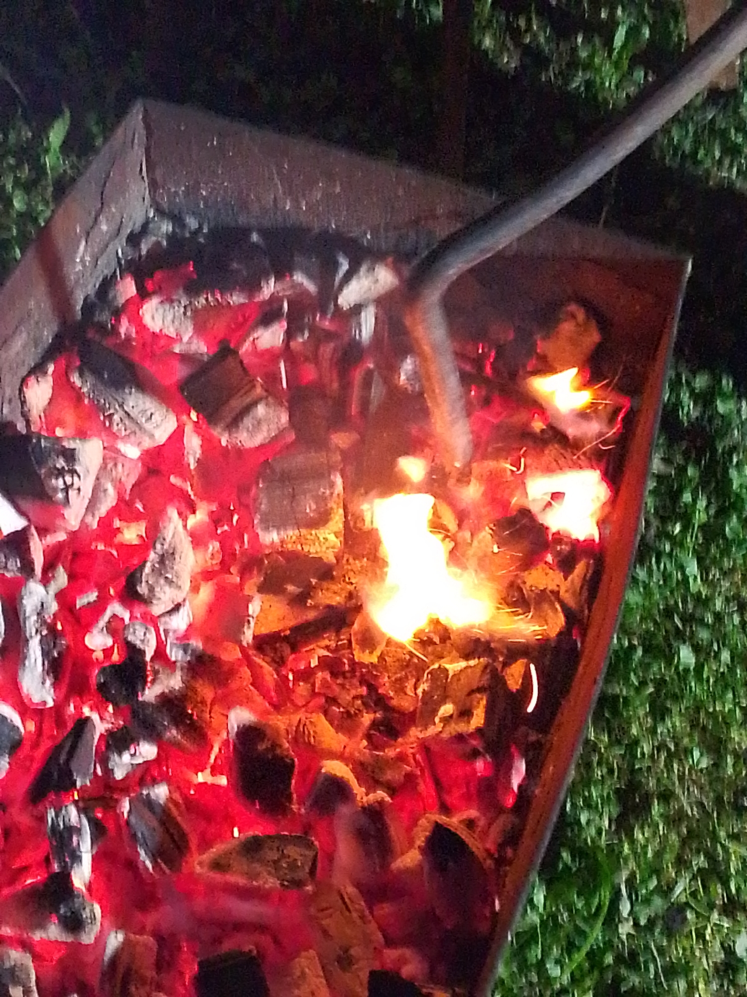 The image shows a close-up view of a burning fire inside a rectangular metal fire pit. The fire consists of glowing embers and flames, with some of the flames visible as bright orange and yellow. A metal rod or tool is inserted into the fire, likely for stoking it. The fire pit sits on a surface that appears to be grass or vegetation. The photo was taken at night. Based on the location provided, the scene is likely set in Прикамский, Russia.