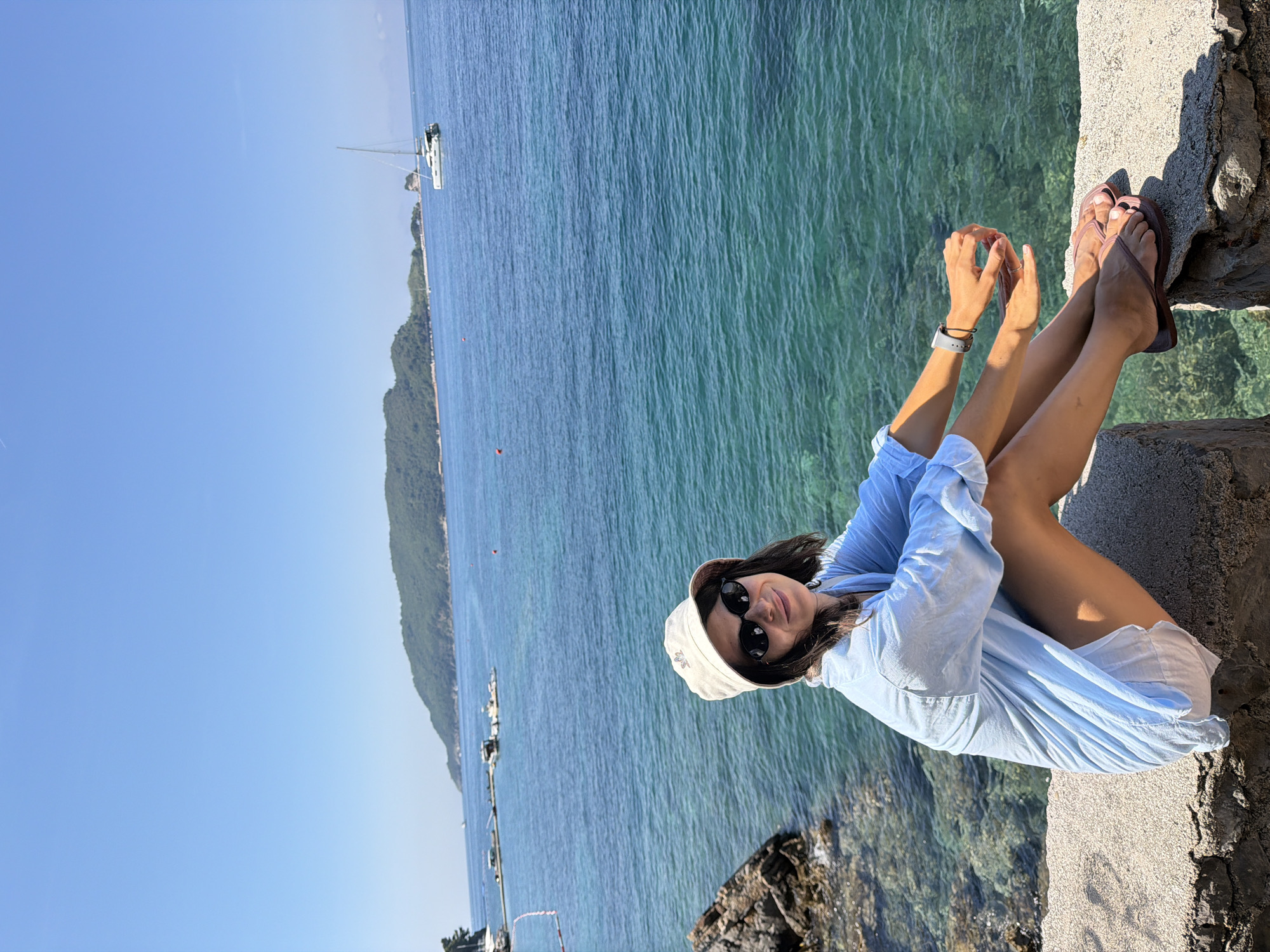 In the city of Boreti, Montenegro, a young woman is pictured sitting on a stone wall overlooking the Adriatic Sea. She is wearing a light blue shirt, white shorts, a beige bucket hat, and sunglasses. The sea is a vibrant turquoise, with a sailboat and several smaller boats visible in the distance. The sky is clear and blue, indicating a sunny day. The woman is looking towards the camera with a slight smile. Her hands are clasped in front of her. The environment appears to be a coastal area with rocky terrain and lush green hills in the background.