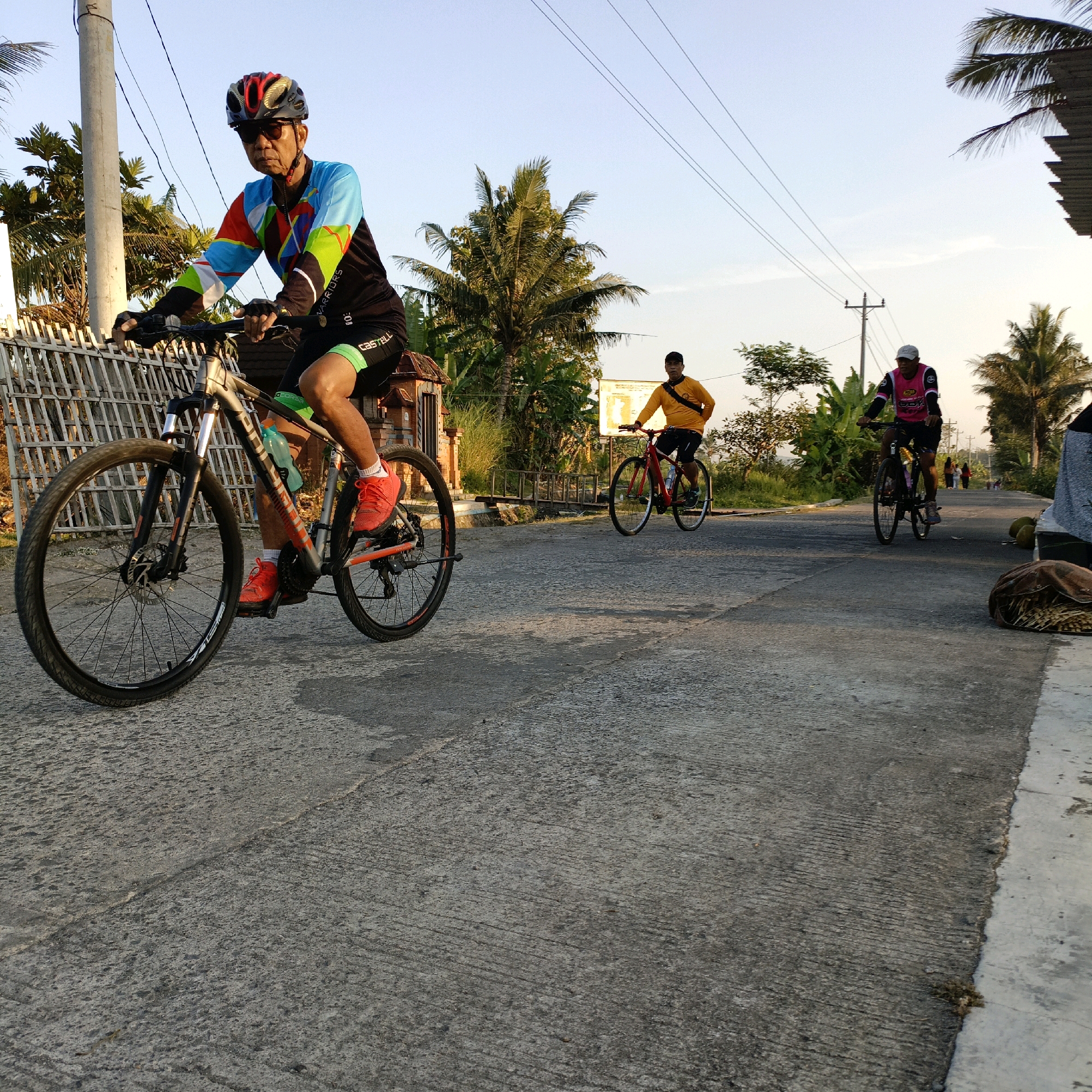 An elderly man in a colorful cycling jersey, black cycling shorts, and a red and black helmet rides a silver mountain bike on a concrete road. He is wearing sunglasses and red cycling shoes. To his right, another man in a yellow shirt and black shorts rides a red bicycle. Further ahead, a person in a pink and black cycling jersey and a white cap rides a bicycle. Palm trees and other vegetation line the road, with power lines overhead. A woven fence is visible to the left of the foreground cyclist. The scene appears to be set in Salatiga, Indonesia, during daylight hours with clear skies.